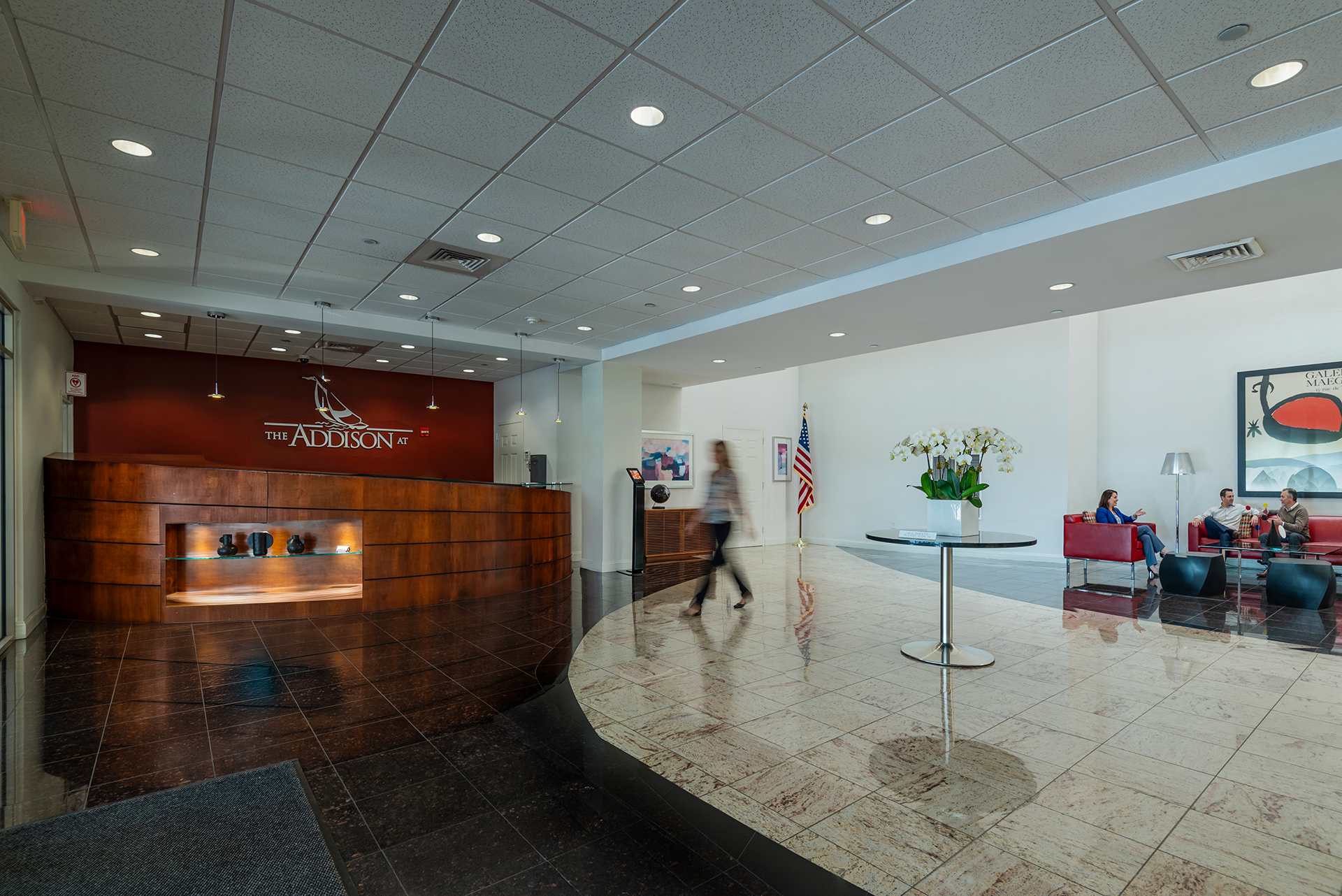 Hotel lobby with a curved wooden reception desk on the left and a seating area on the right screened by a glass partition. The seating area has red couches, a black coffee table, and a tall floor lamp, with people sitting and talking. A large flowerp