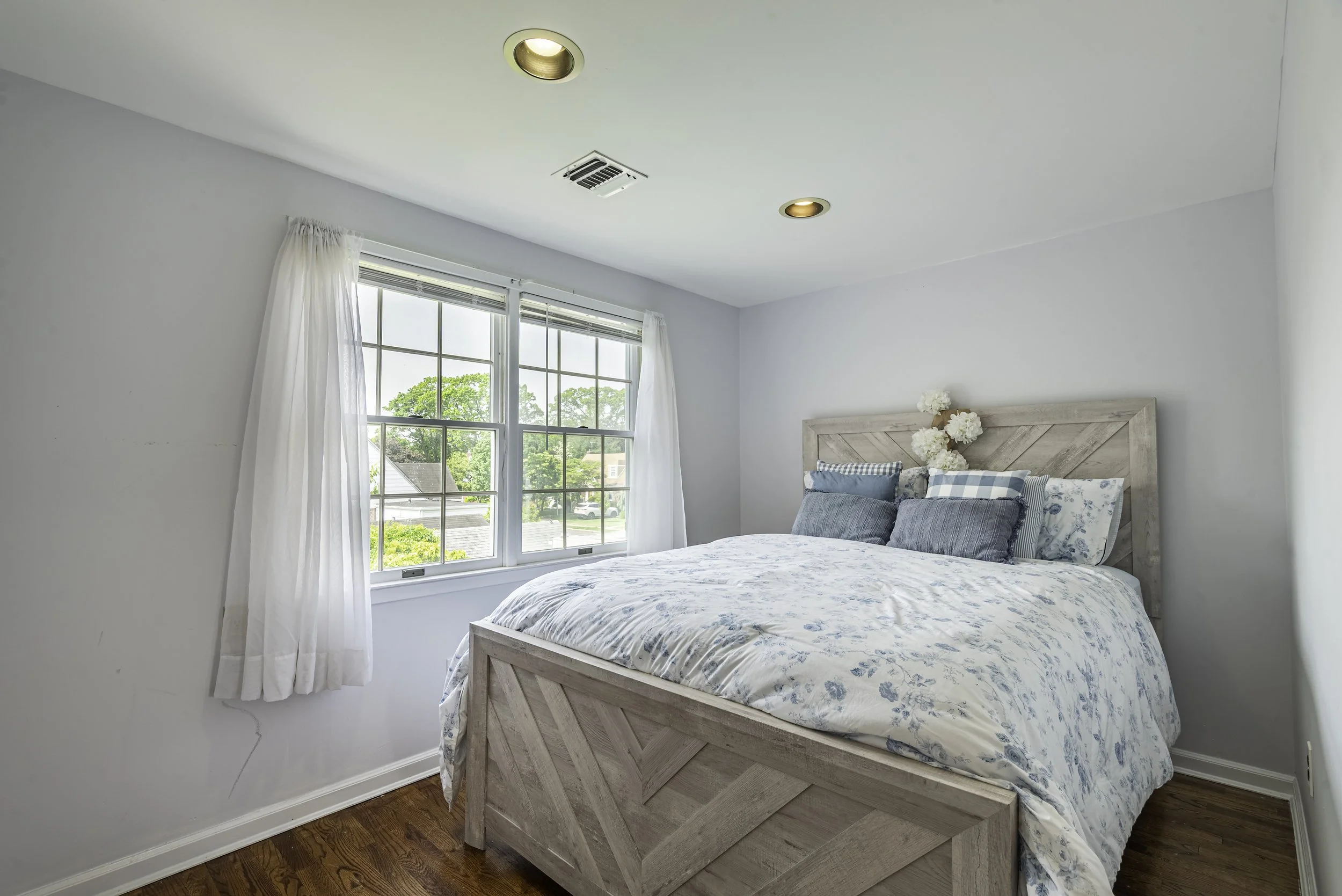 Bright bedroom with hardwood floors, large window, recessed lighting, modern wood bed frame, and soft natural light filling the space.