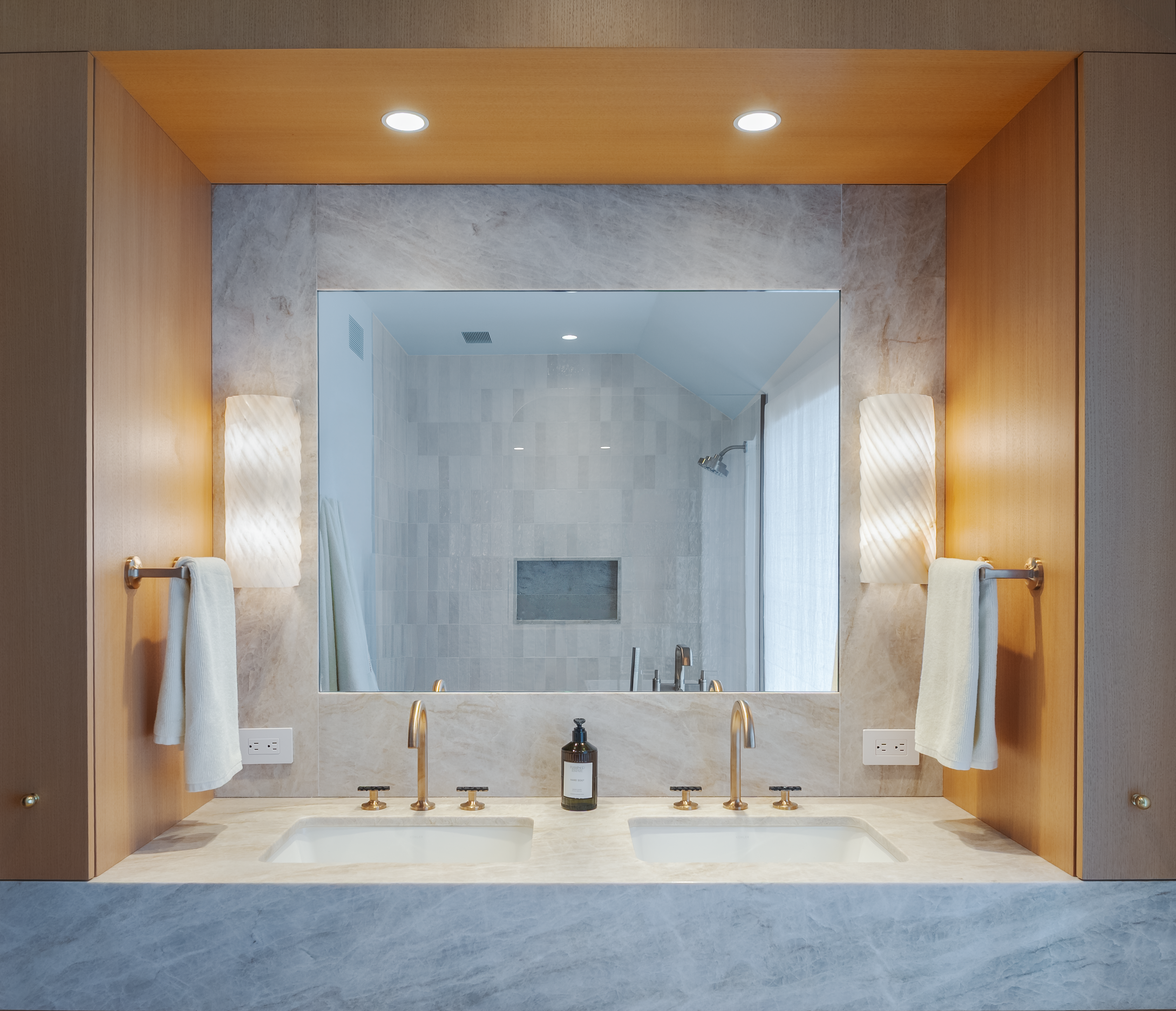 alt text
Double bathroom vanity with brass faucets, pale stone countertop, warm wood surround, and sculptural wall sconces flanking a large mirror.