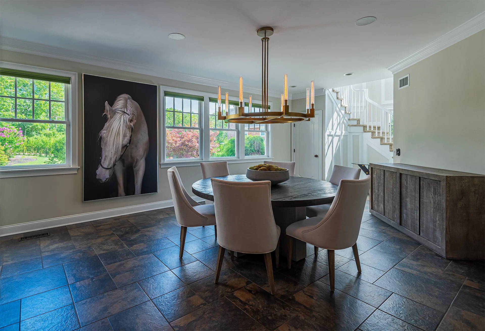 Dining room with round wooden table surrounded by six beige upholstered chairs. On the wall is a large portrait of a white horse. Natural light from multiple windows reveals a view of lush green trees outside. A modern chandelier hangs above the tabl