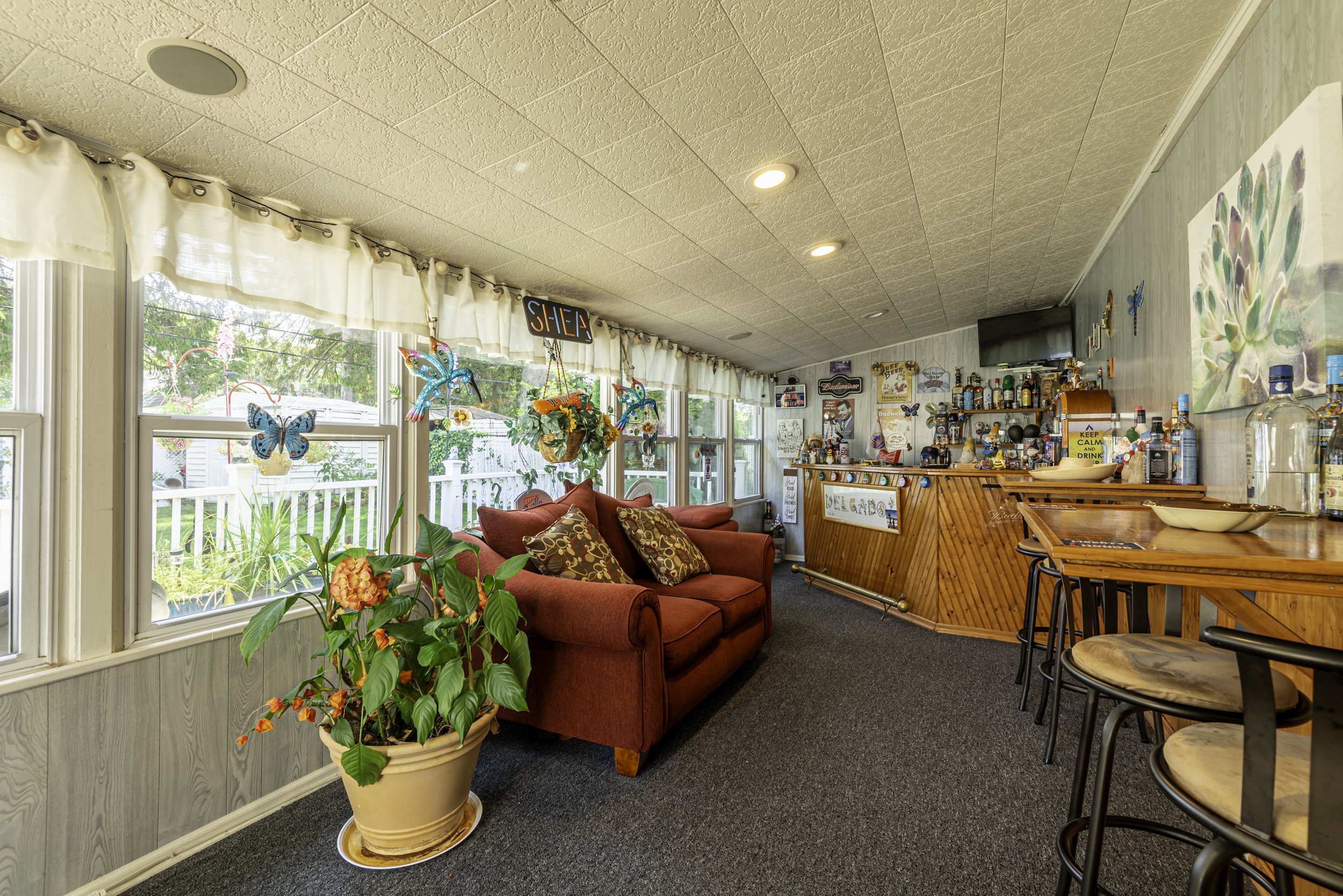 Bright enclosed sunroom with bar area, cozy seating, decorative wall accents, and large windows overlooking the backyard.
