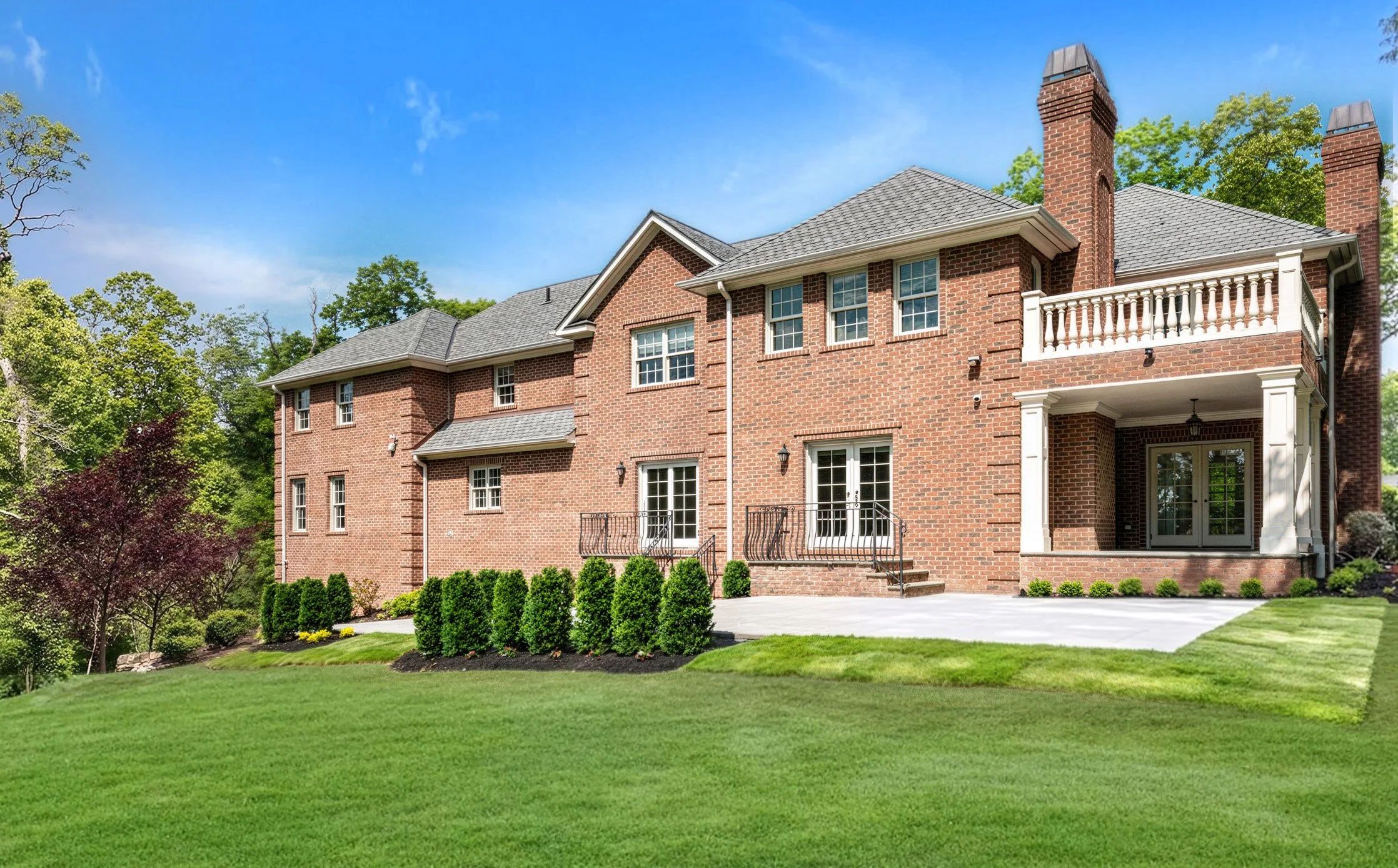 Rear exterior of a custom brick colonial estate featuring a covered balcony, wrought iron railings, French doors, and
  professional landscaping — photographed by Palma Design. Architectural photography for luxury homebuilders and designers in
  New 