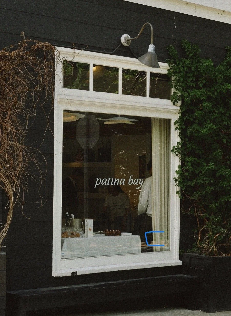 A window with the words 'patina bay' written on it, part of the exterior of a building with black walls, surrounded by green ivy and a dry vine, with a sidewalk beneath.