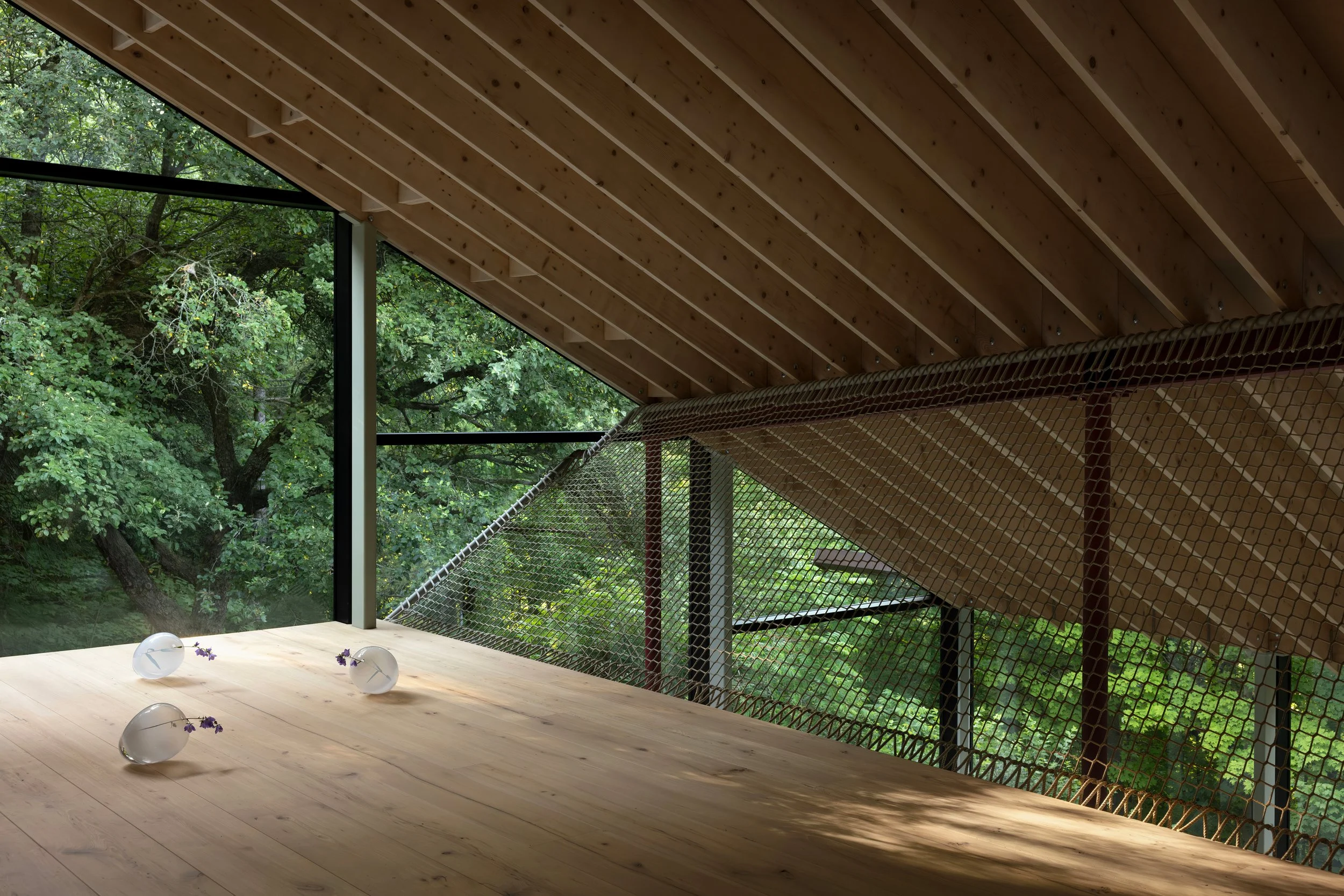 Interior view of a modern building with a wooden ceiling, large glass windows, and a forested landscape outside. There are small glass vases with purple flowers on a wooden table.