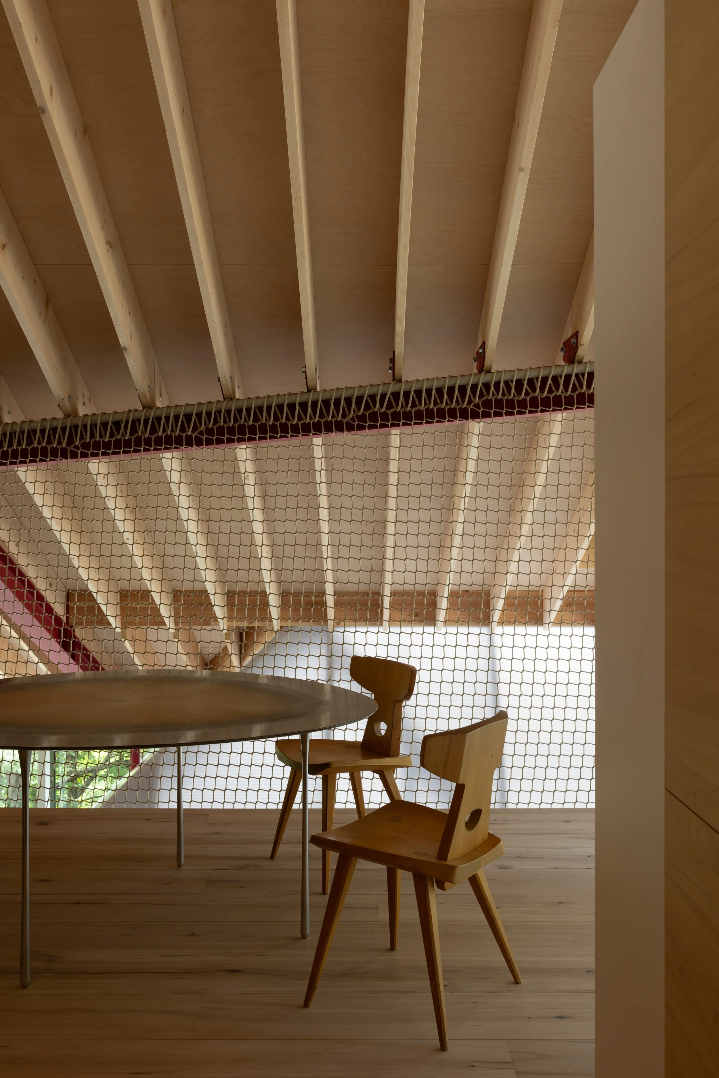 Interior view of a dining area with a round wooden table, wooden chairs, and a partial view of a loft or upper level with exposed wooden beams and a netted barrier.