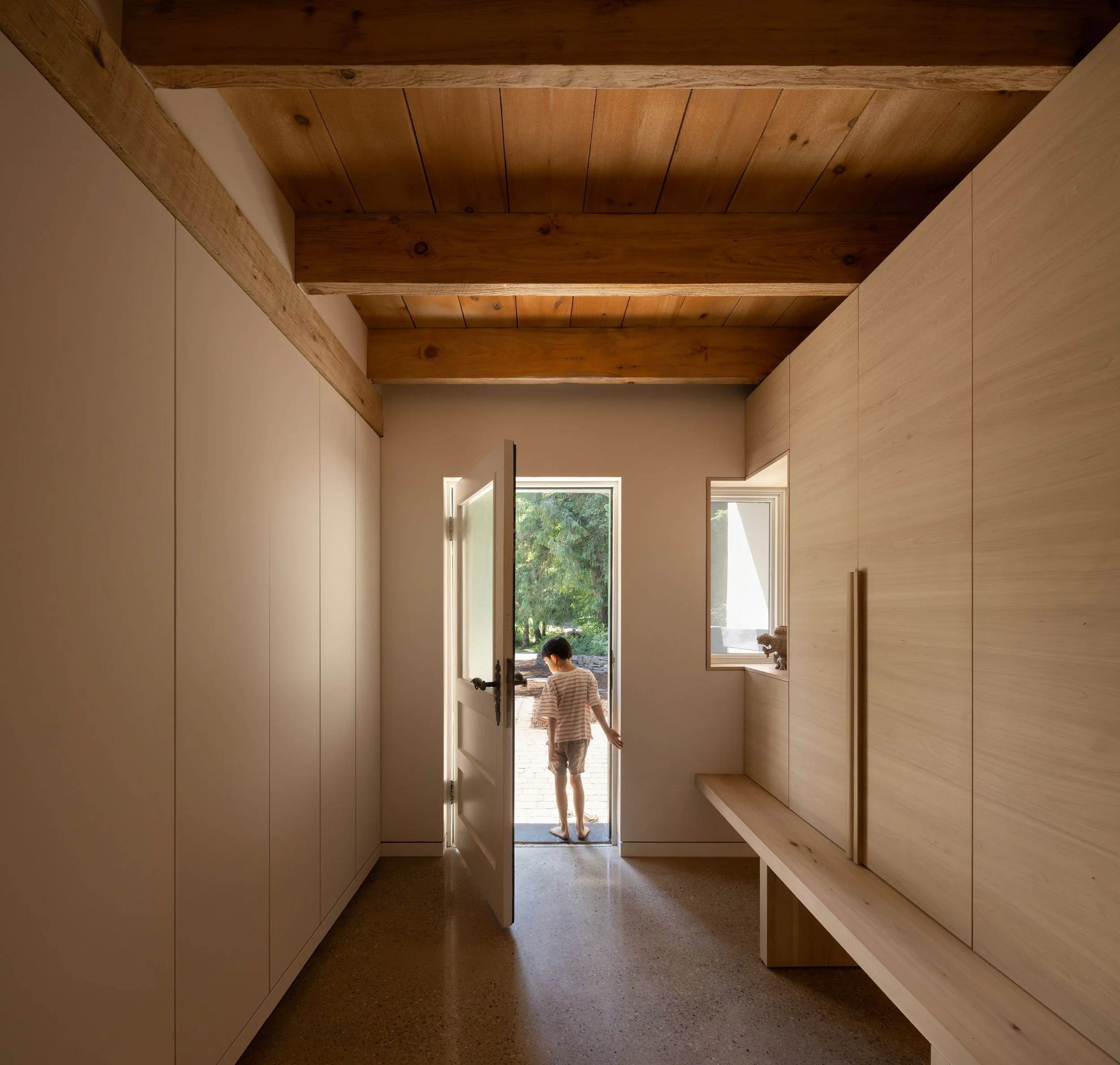 A person walking through a doorway from a room with wooden shelves and a wooden ceiling, into a brightly lit outdoor area with trees visible outside.
