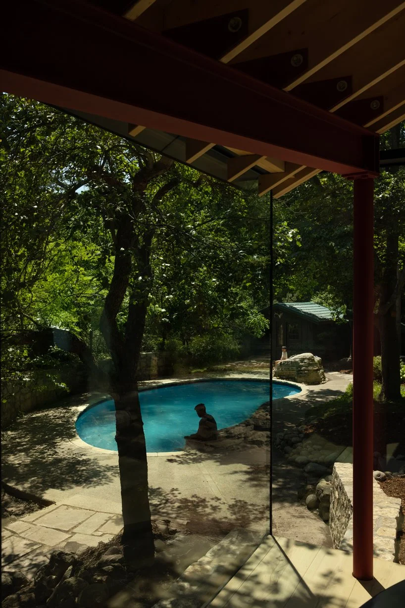 View through a glass window showing a backyard with a blue kidney-shaped pool, trees providing shade, and a person sitting on the pool edge.