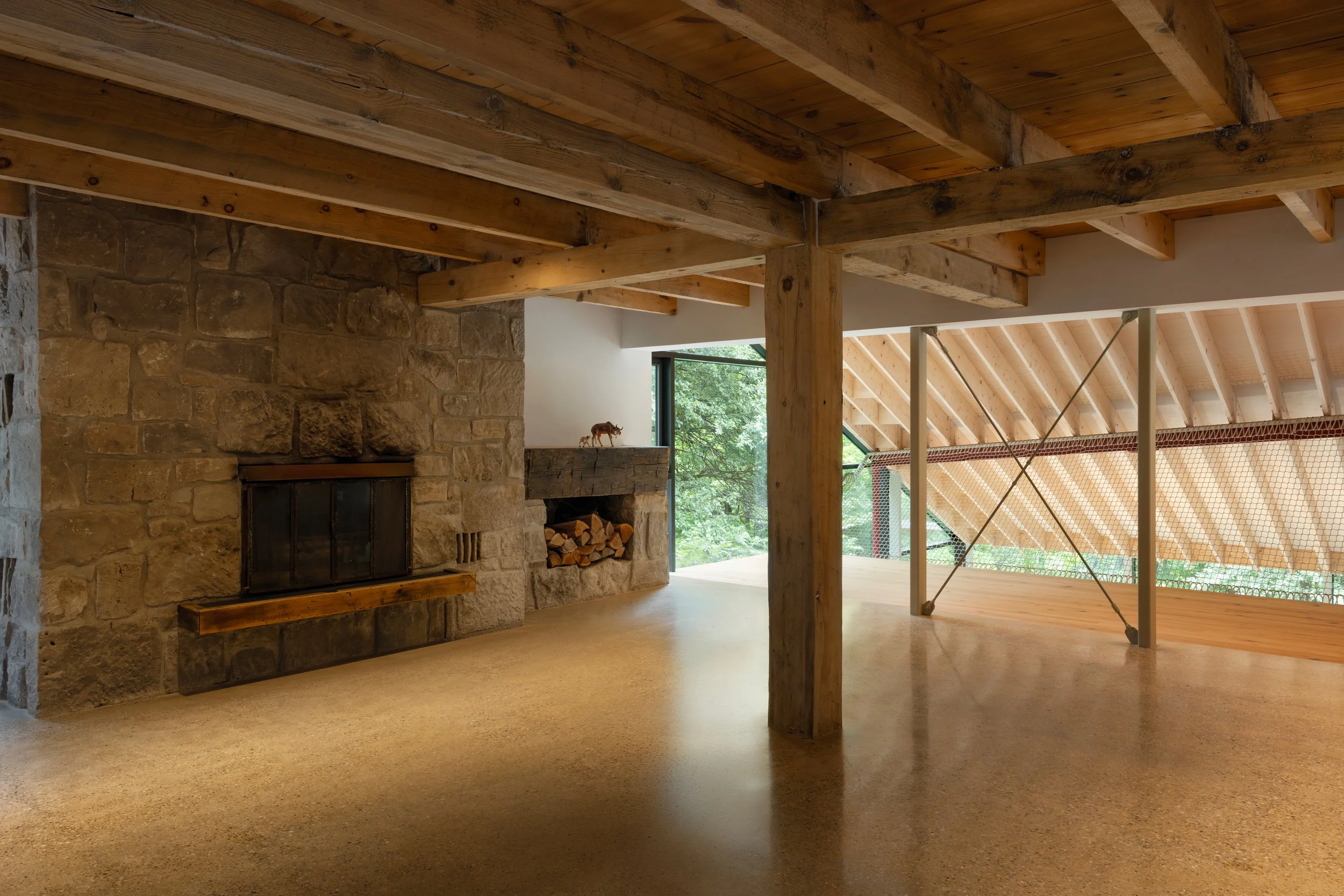 Interior of a modern building with wooden beams and ceiling, stone fireplace, and large windows showing greenery outside.