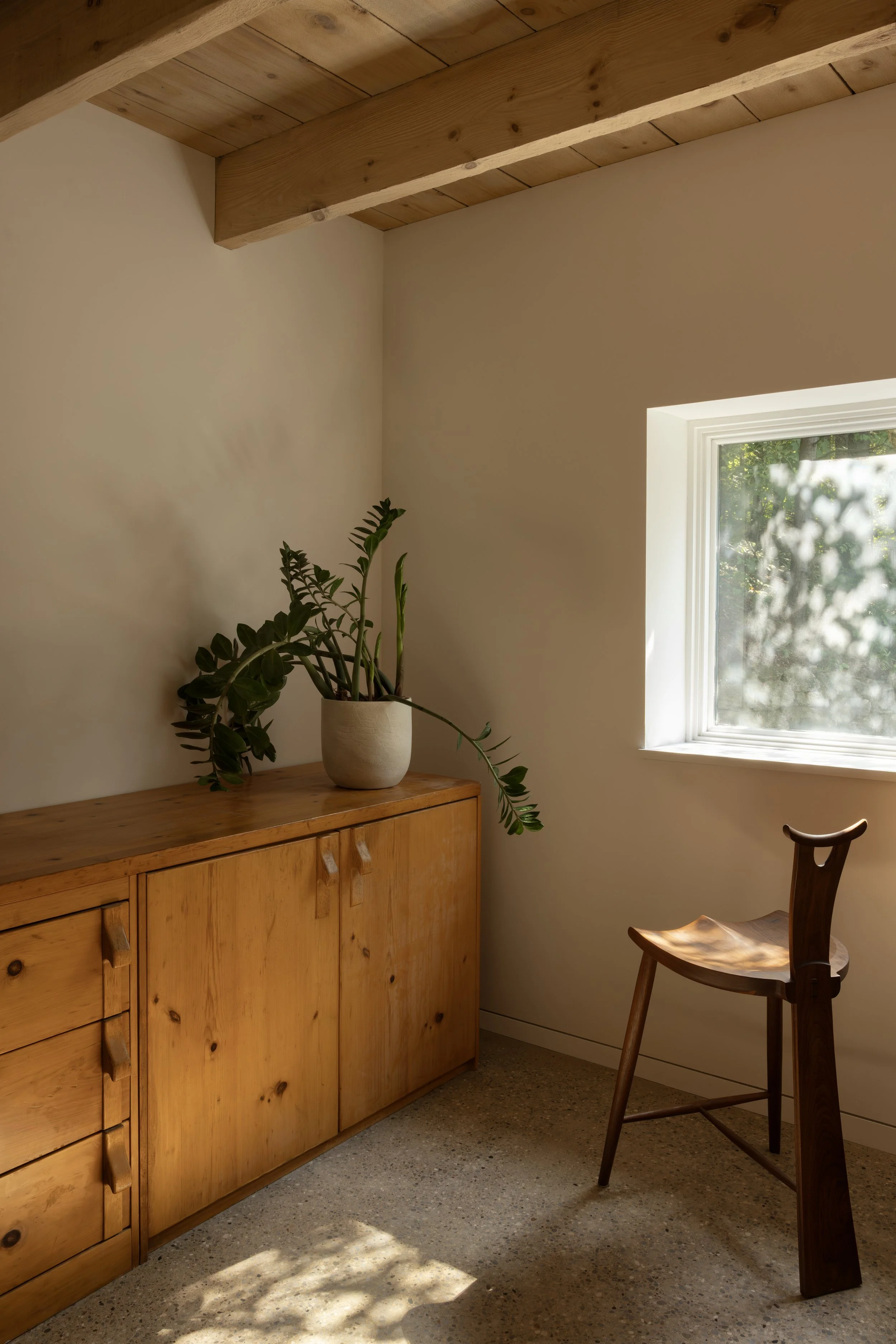 Interior room with a wooden cabinet, potted plant on top, a window with sunlight and greenery outside, and a wooden chair with curved backrest.