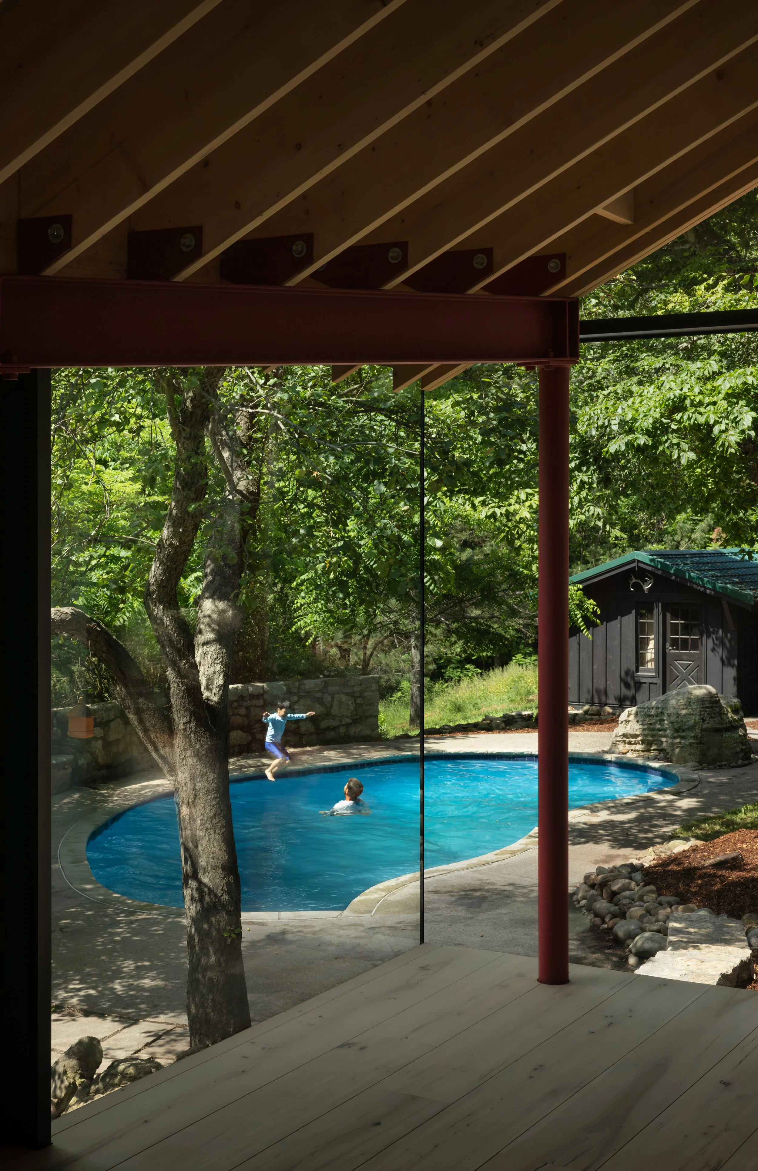Children playing in a backyard swimming pool, viewed from an enclosed porch with wooden roof and support beams, trees and a small black shed in the background.