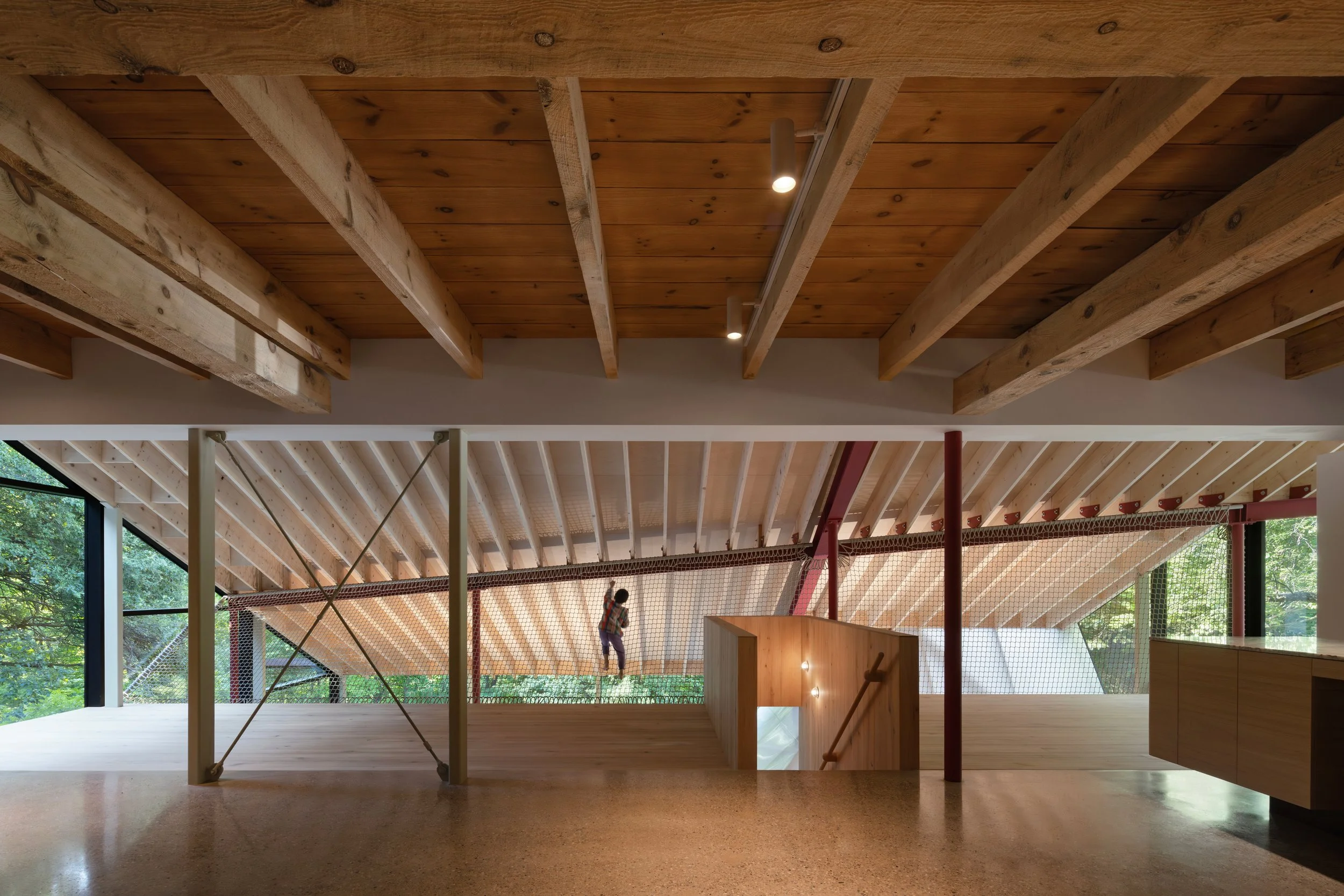 Interior view of a structure with a wooden ceiling, beams, and large windows, showing a person playing badminton outside through the windows.