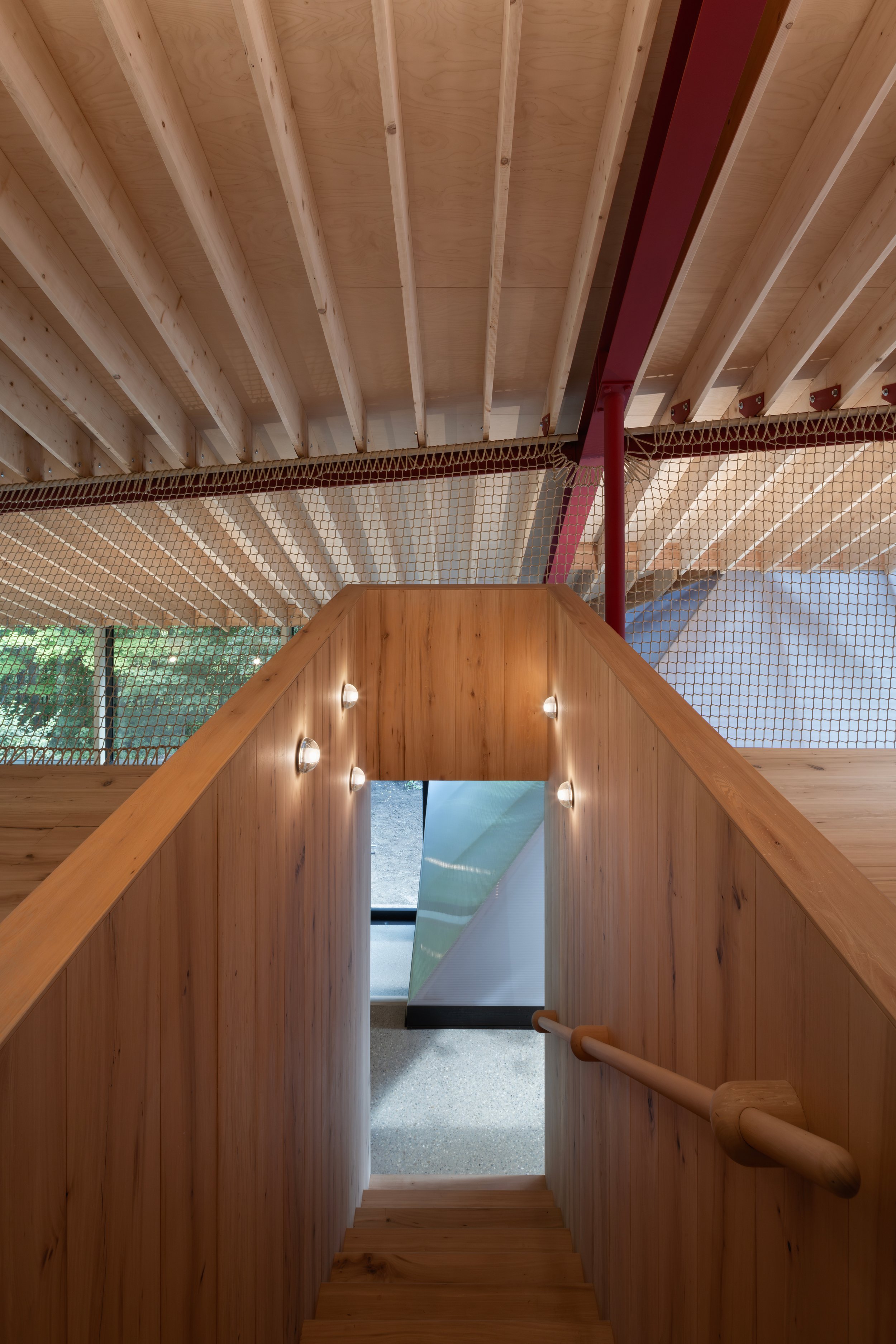 Wooden staircase leading down, with wood-paneled walls, a handrail, and a view of a glass door or window at the bottom, in an interior setting.