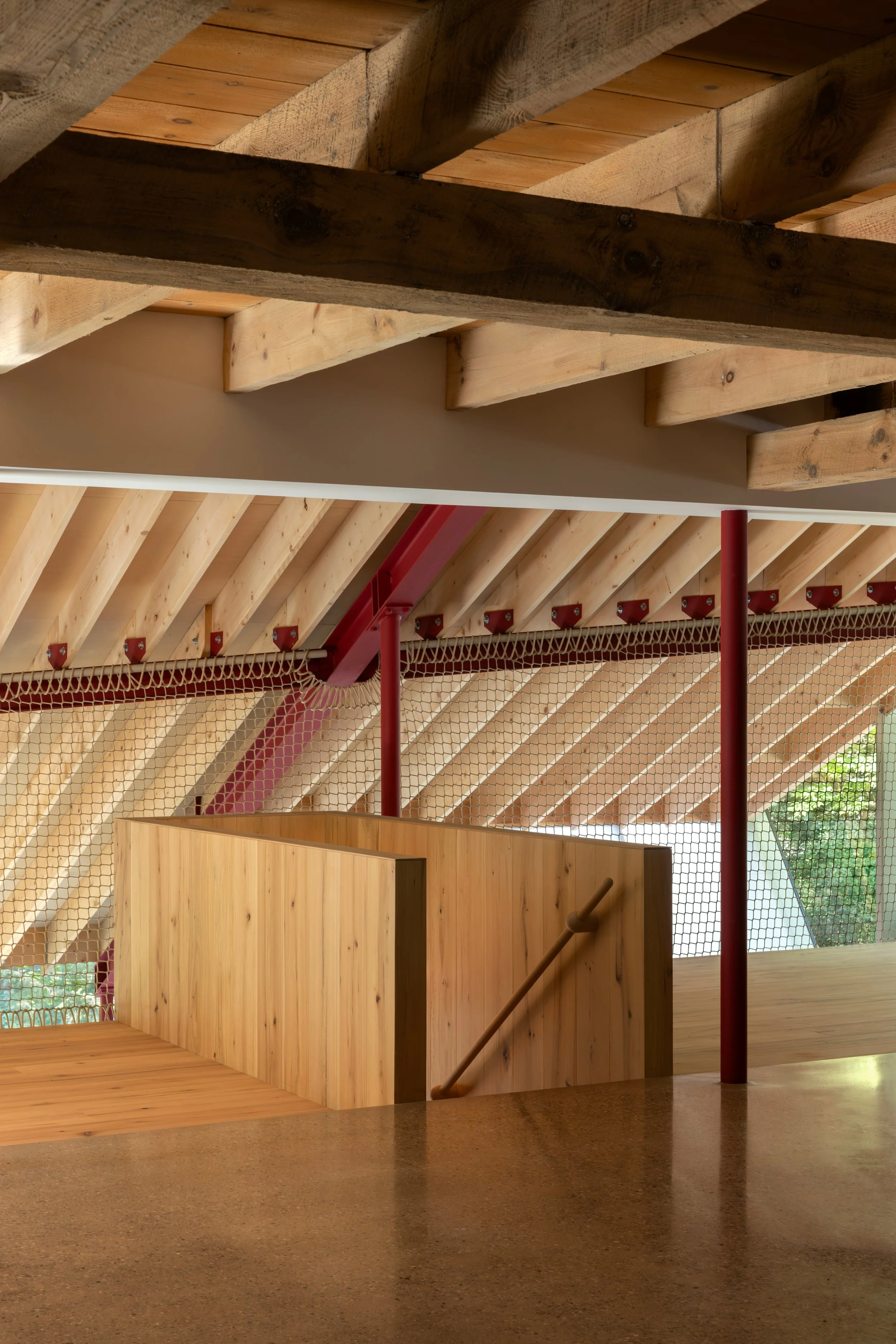 Interior view of a building with wooden ceiling beams and a wooden staircase with railing, yellow wood paneling, and metal framework with mesh netting.