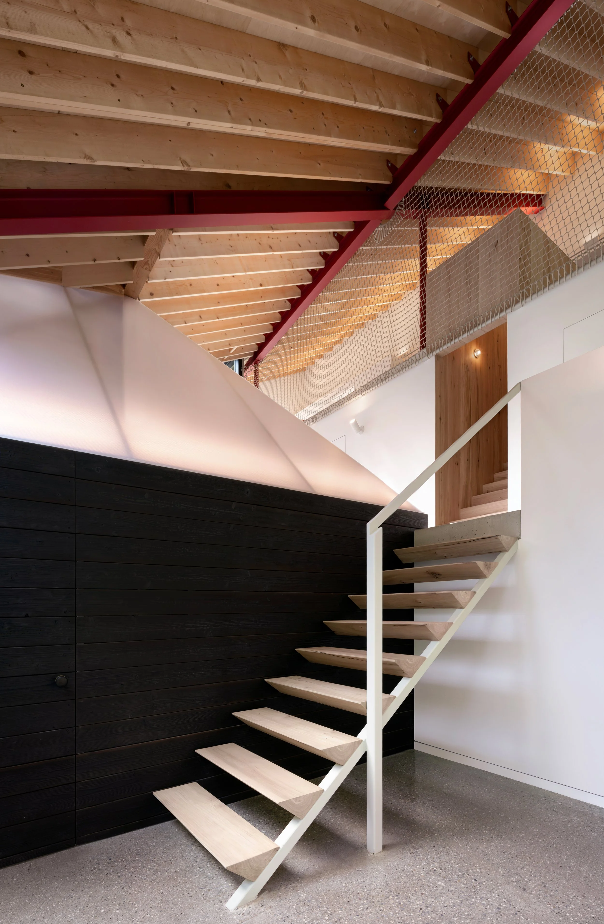 Interior view of a modern staircase with wooden steps, white railing, a wood wall, and a metal mesh railing on the upper level.