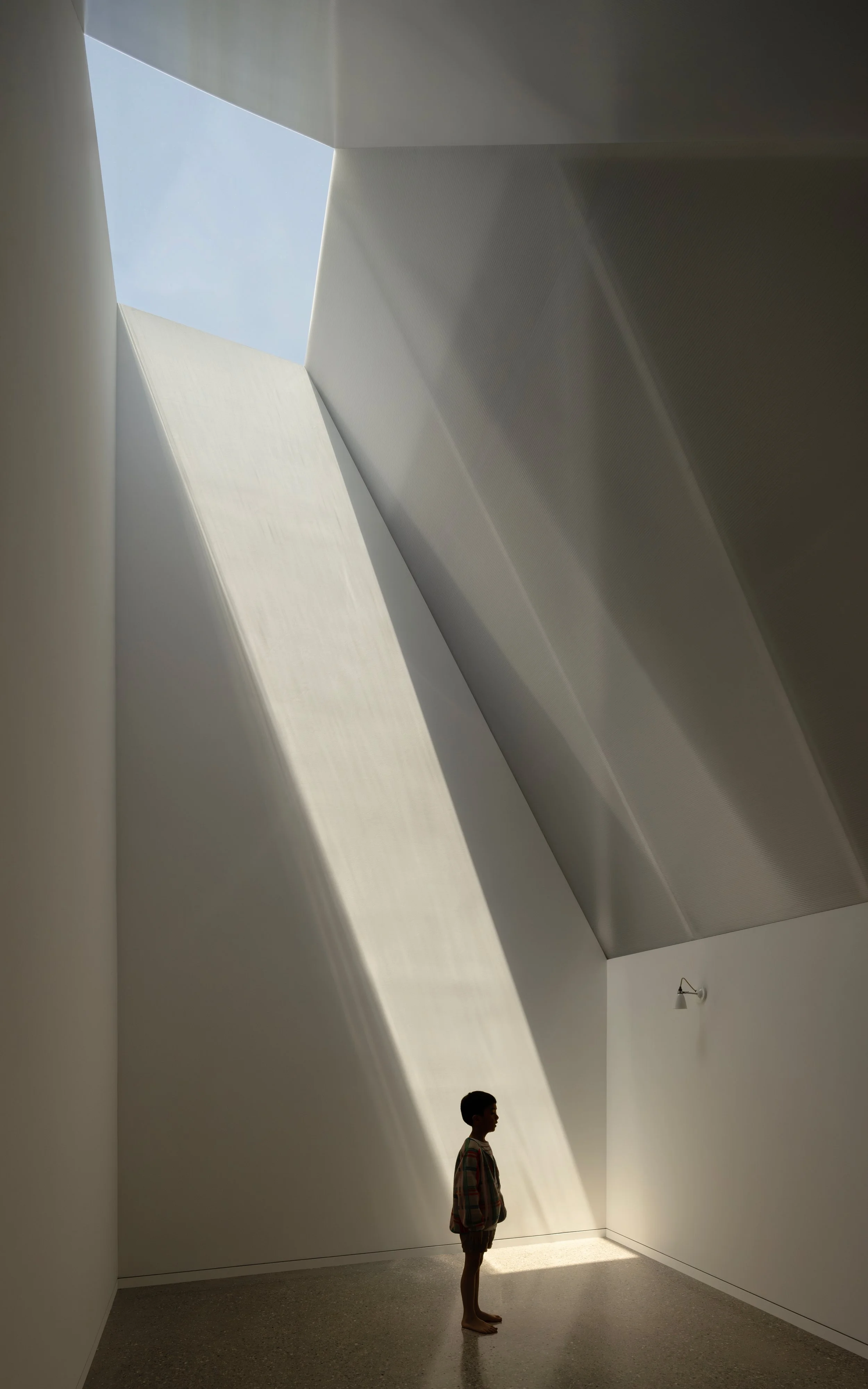 A boy standing barefoot in a minimalist room with sloped white walls and a large skylight, sunlight streaming through and casting shadows.