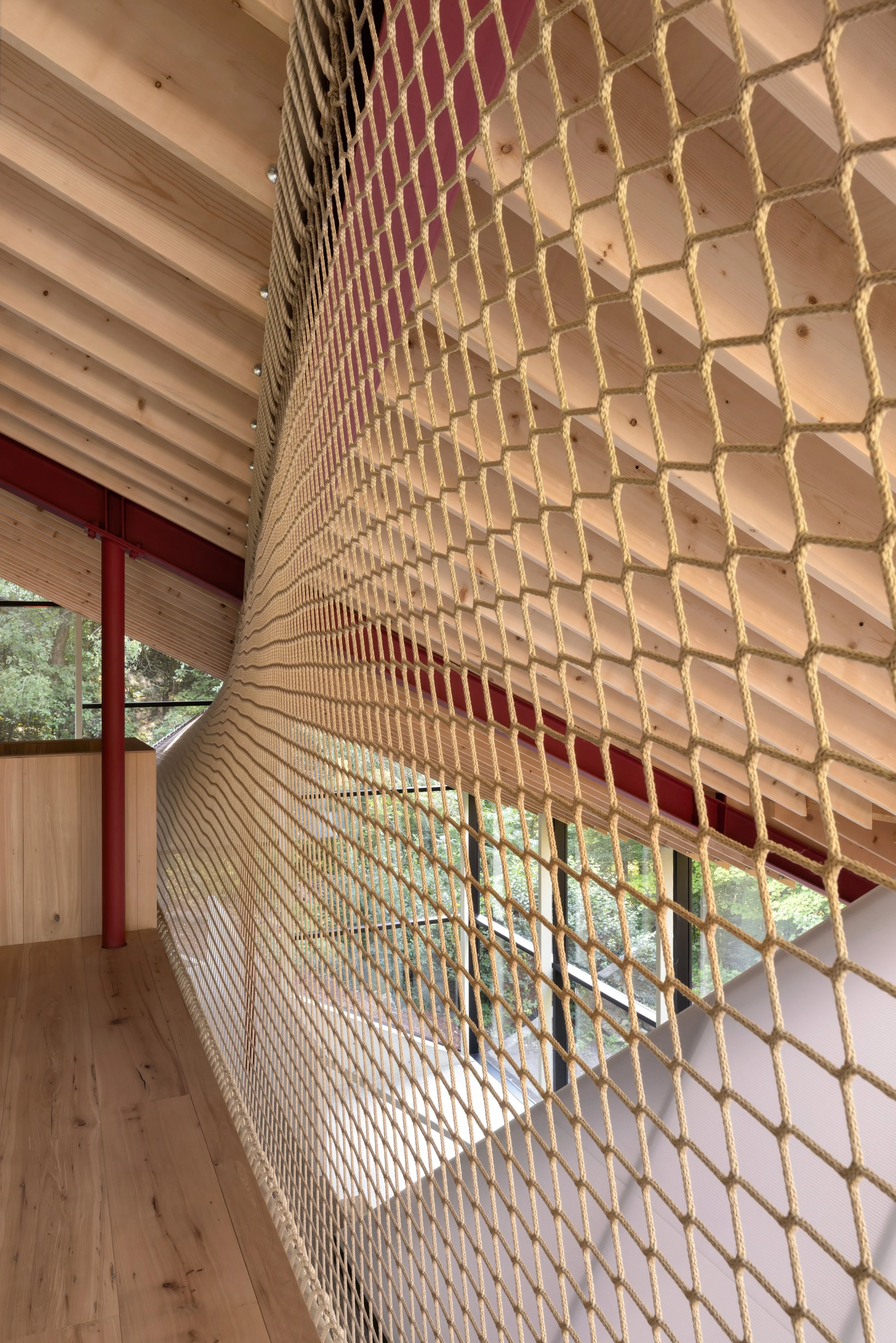 Interior space featuring a wooden ceiling and floor, large windows overlooking a wooded area, with a beige rope netting barrier.