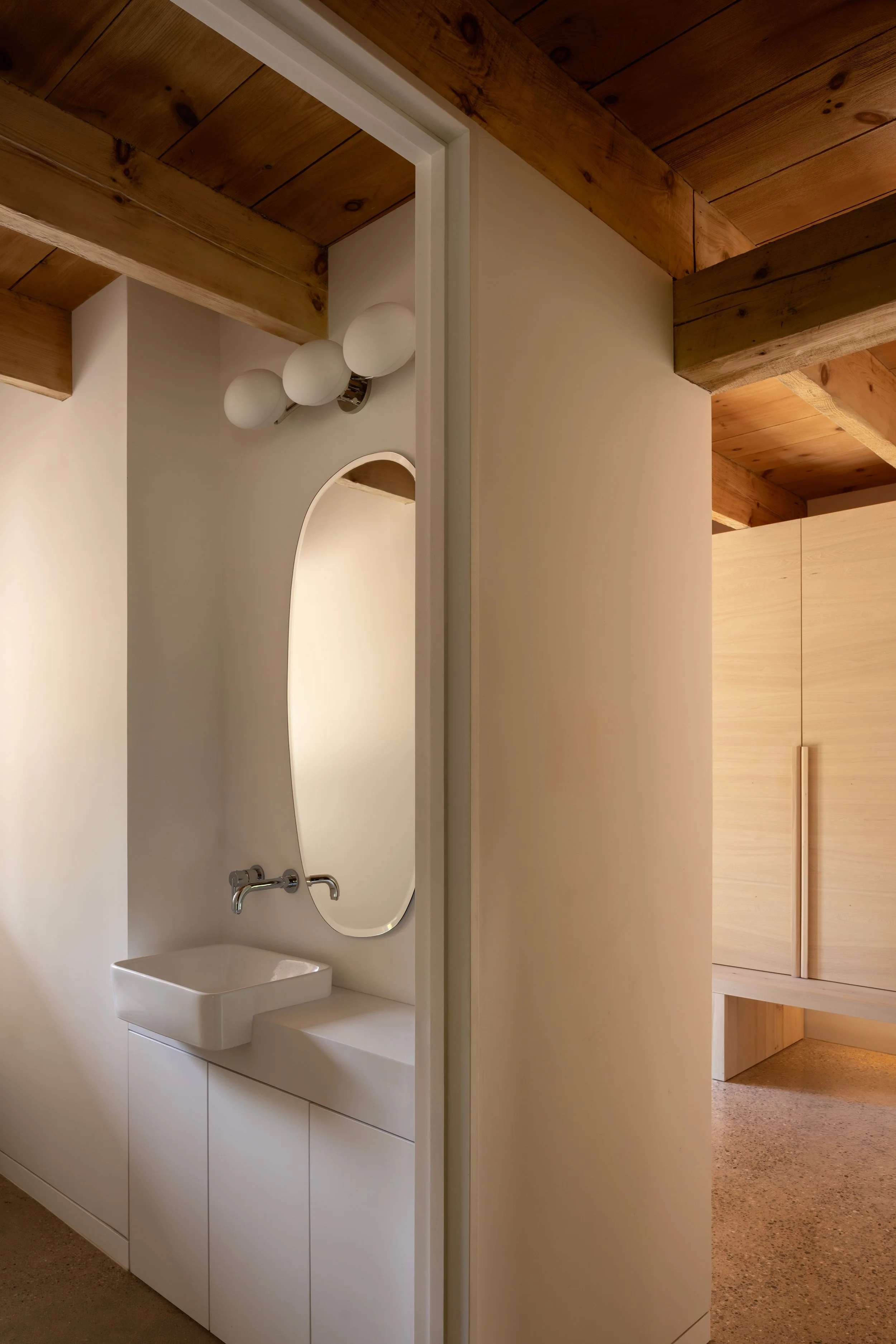 Bathroom with a white cabinet, a sink, an oval mirror, and three globe-shaped light bulbs on the wall, with a wooden ceiling and flooring in the background.