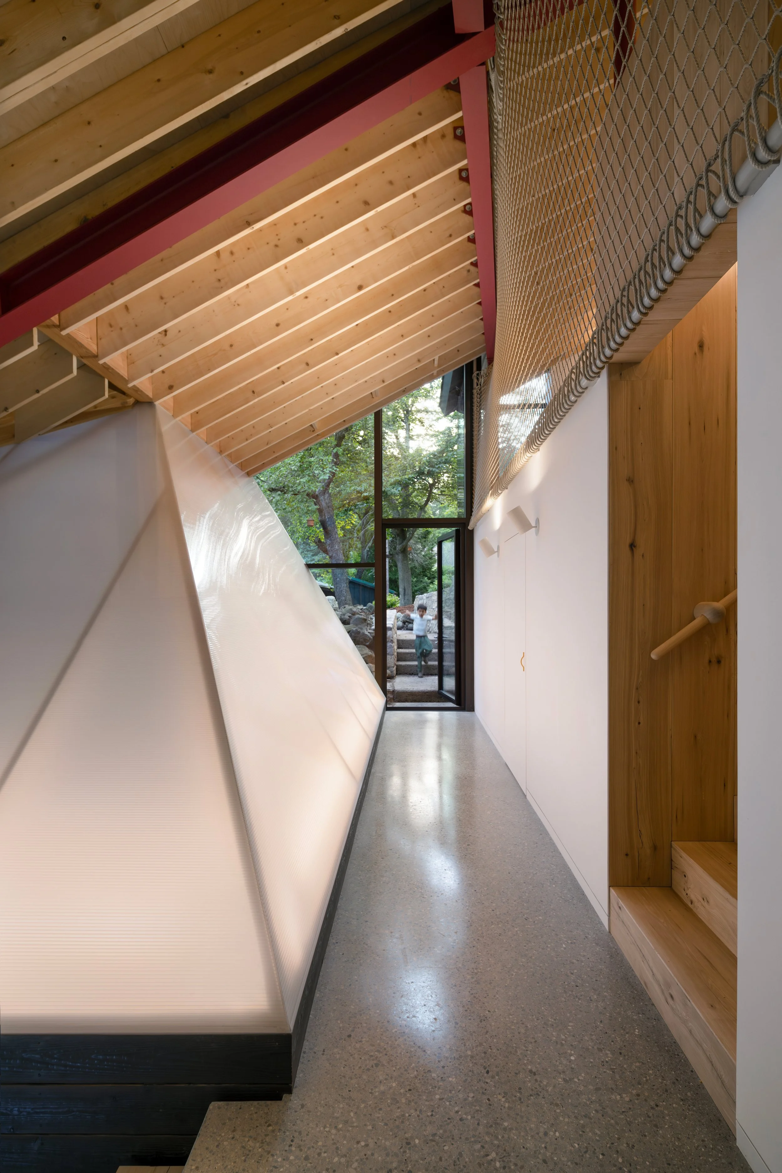 Indoor hallway with wooden ceiling and large window showing outdoor trees. On the left, a large white object possibly part of a slide or wall. On the right, wooden and white wall with a built-in wooden shelf. The floor is concrete, and a person can b