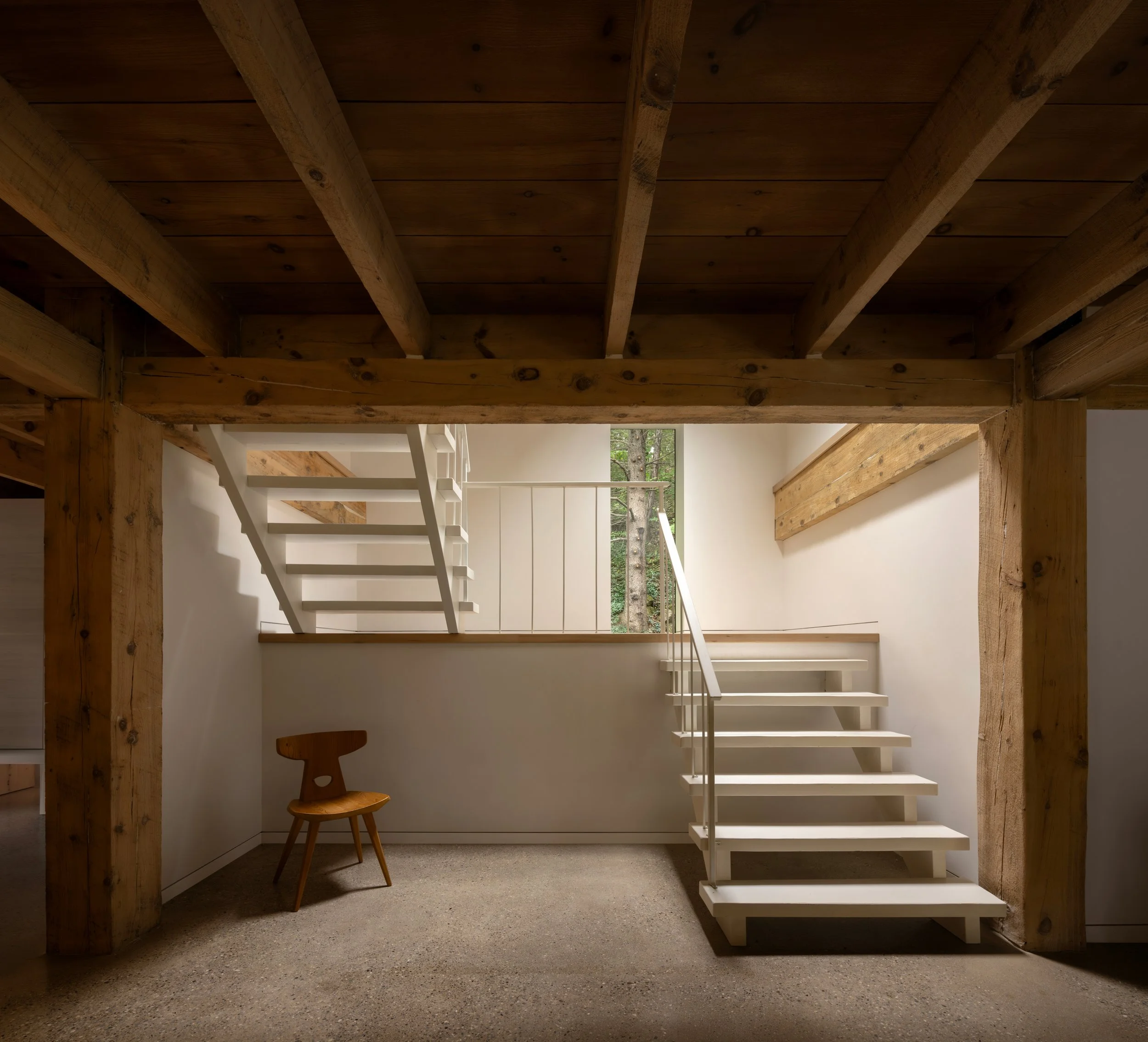 Interior space with a wooden staircase and a wooden chair, a window with a view of trees, and wooden beams on the ceiling.