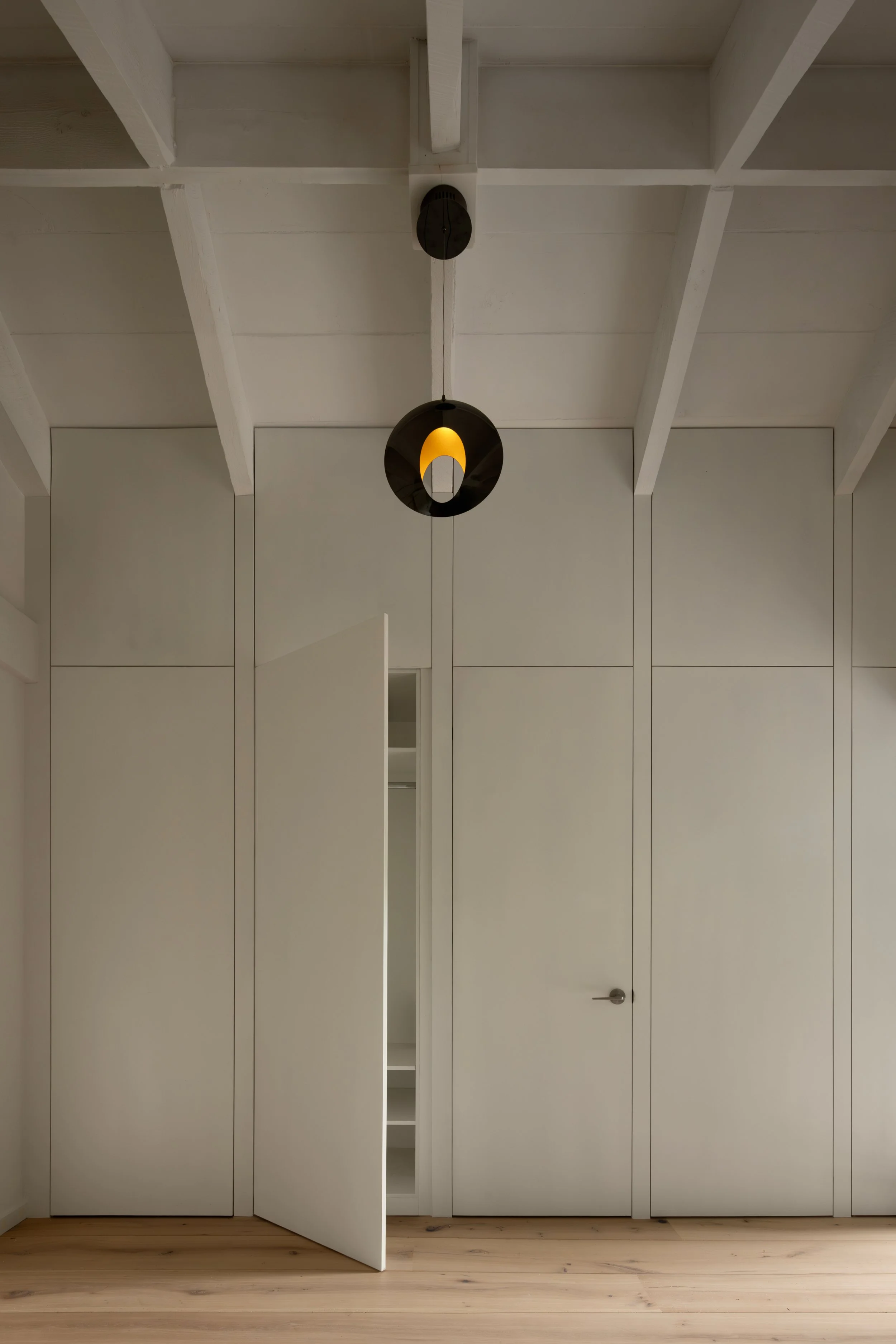 White built-in closet with many closed doors, an open door revealing shelves, in a room with light wood floors and a white ceiling with wooden beams. A modern black pendant light hangs from the ceiling.