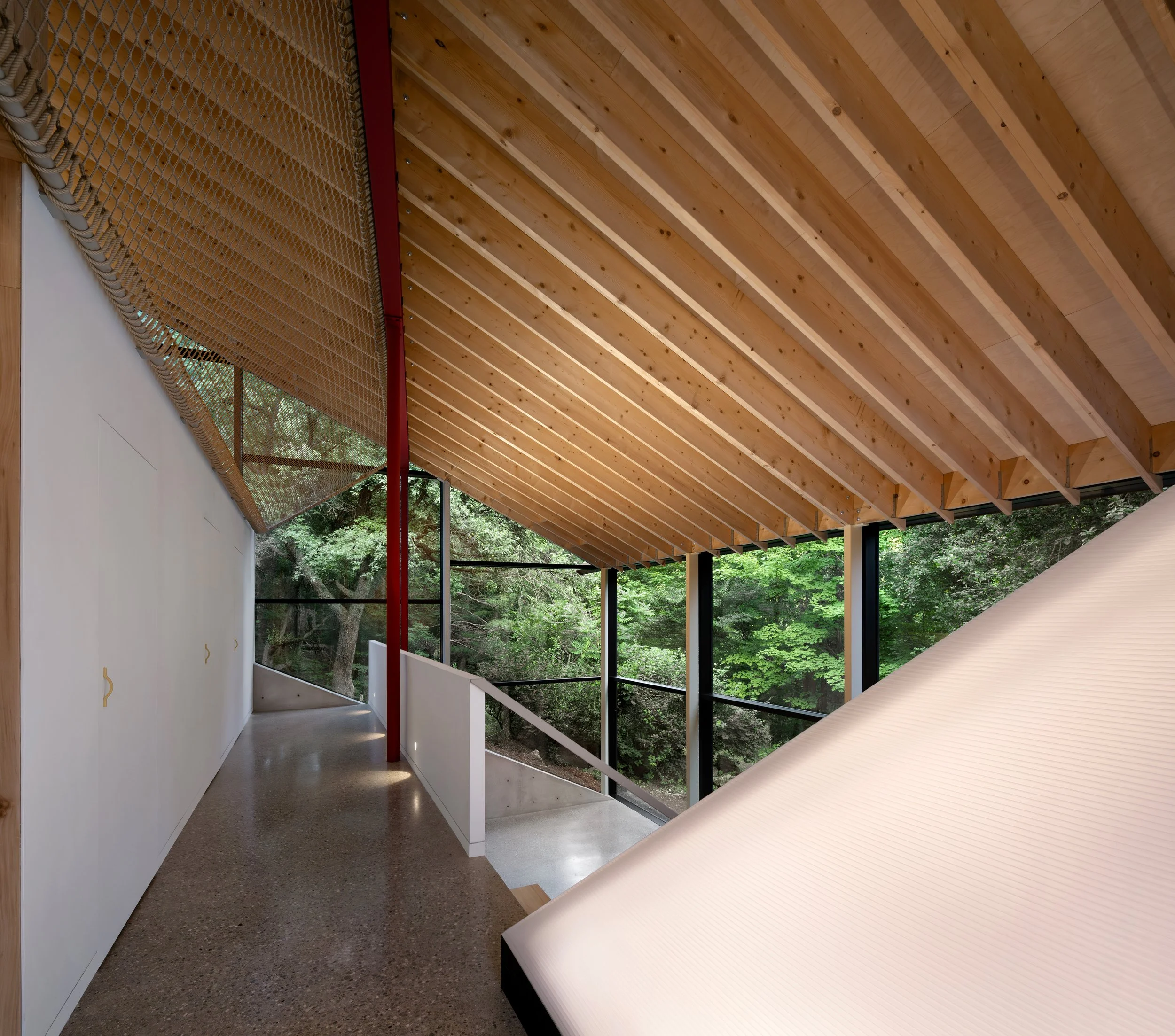 Interior space with a curved wooden ceiling, a white wall with hooks, a netted barrier, a window showing green trees outside, and a beige table.