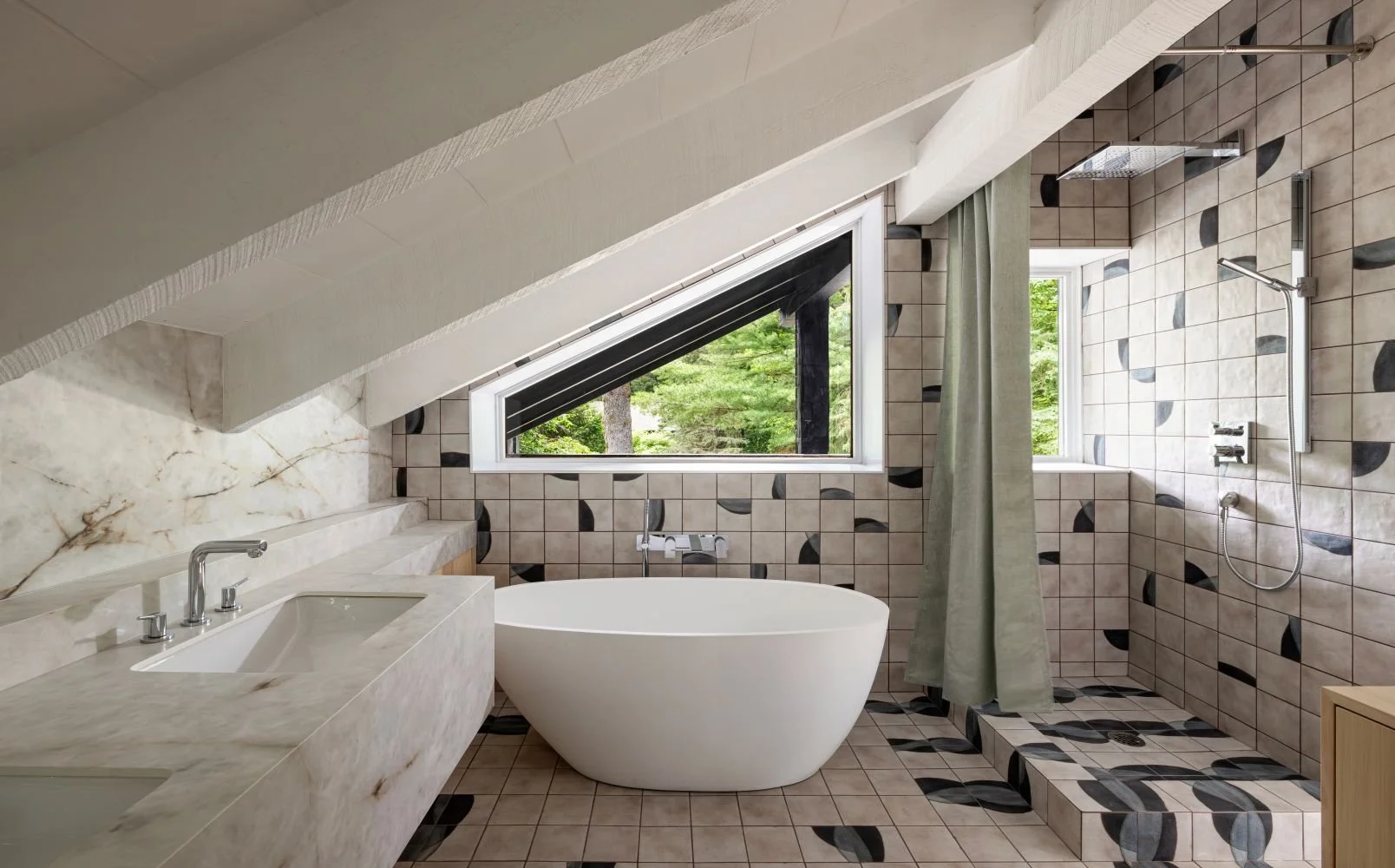 Modern bathroom with sloped ceiling, marble countertop with sink, free-standing bathtub, tiled walls with black and gray accents, shower area with rain shower and handheld shower, window showing greenery outside, beige and black patterned floor tiles
