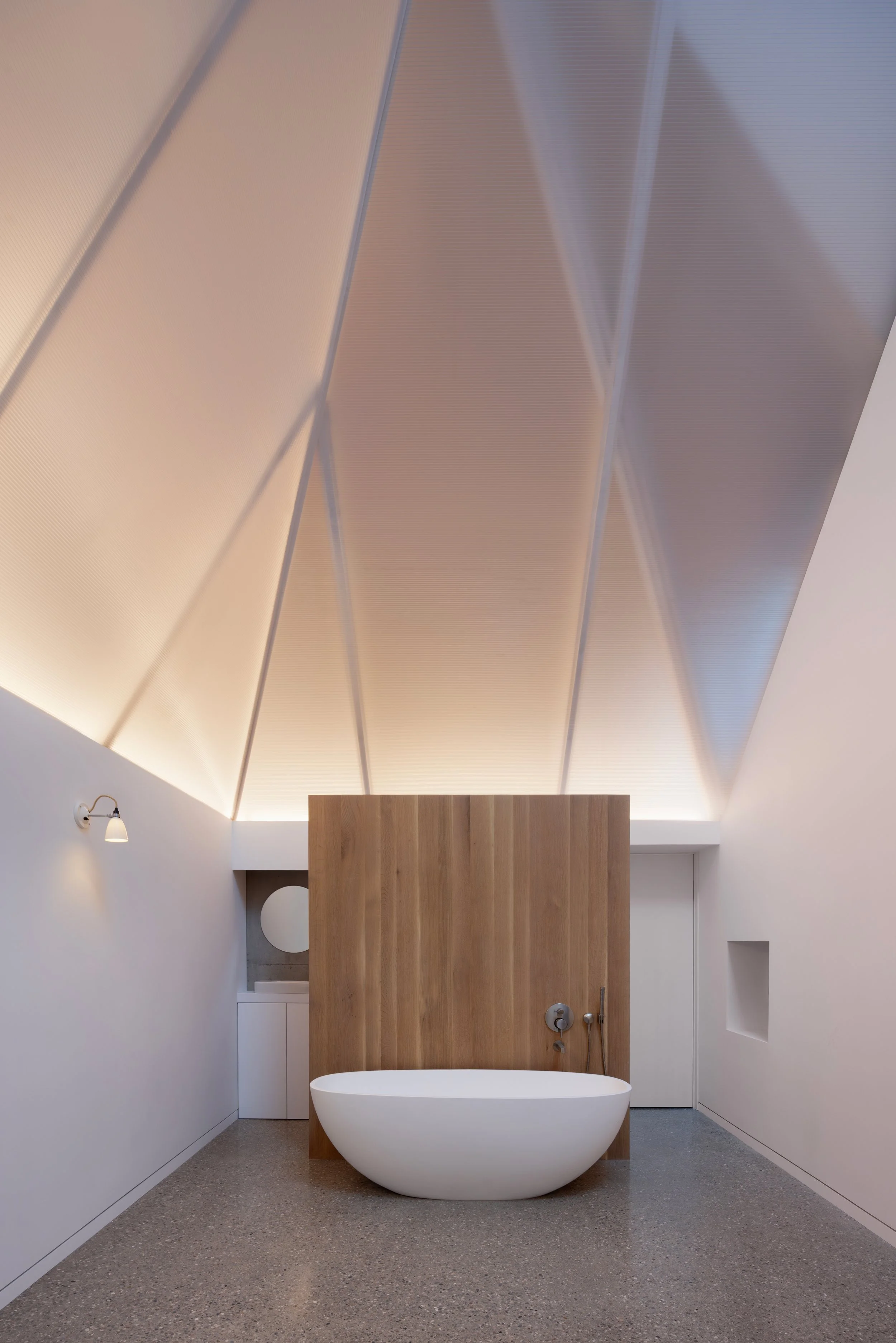 Modern minimalist bathroom with a freestanding bathtub, tall wooden partition, sloped ceiling with acoustic panels, and simple wall-mounted light fixtures.