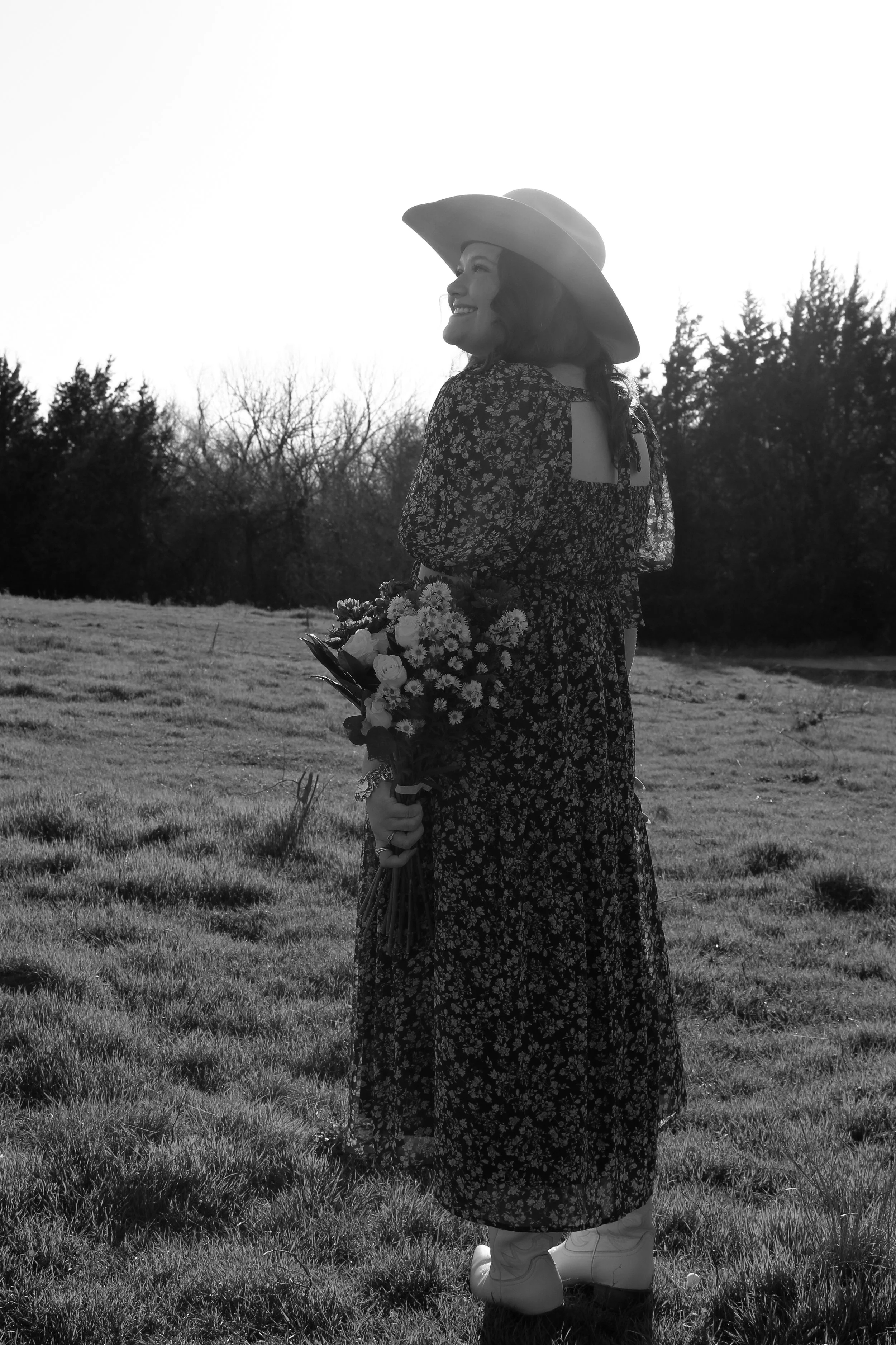 A woman in a floral dress and cowboy hat standing in a grassy field, holding a bouquet of flowers, smiling and looking to her left.