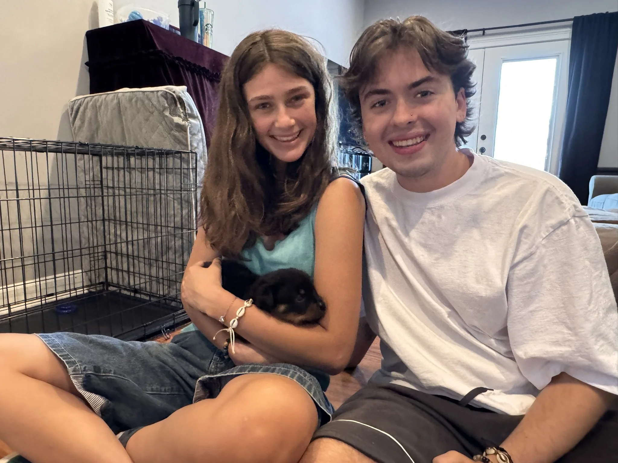 Two young people, a girl with long, wavy brown hair wearing a teal top and a boy with wavy light brown hair wearing a white shirt, smiling and hugging, sitting inside a room near a metal cage, with a door and window in the background.