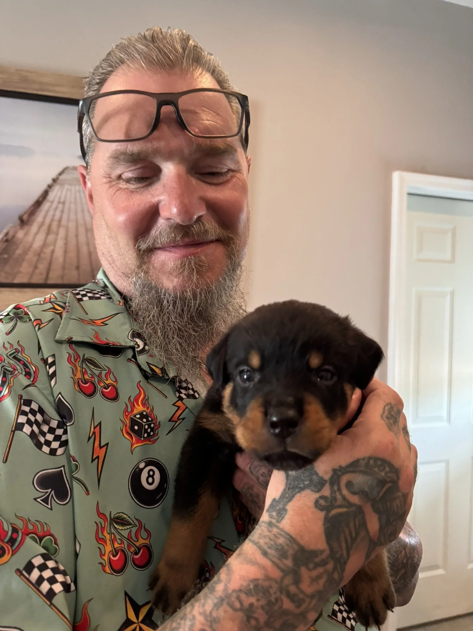 A man with glasses and a beard, featuring tattoos on his arms, is holding a black and tan puppy. The background includes a picture frame and a white door.
