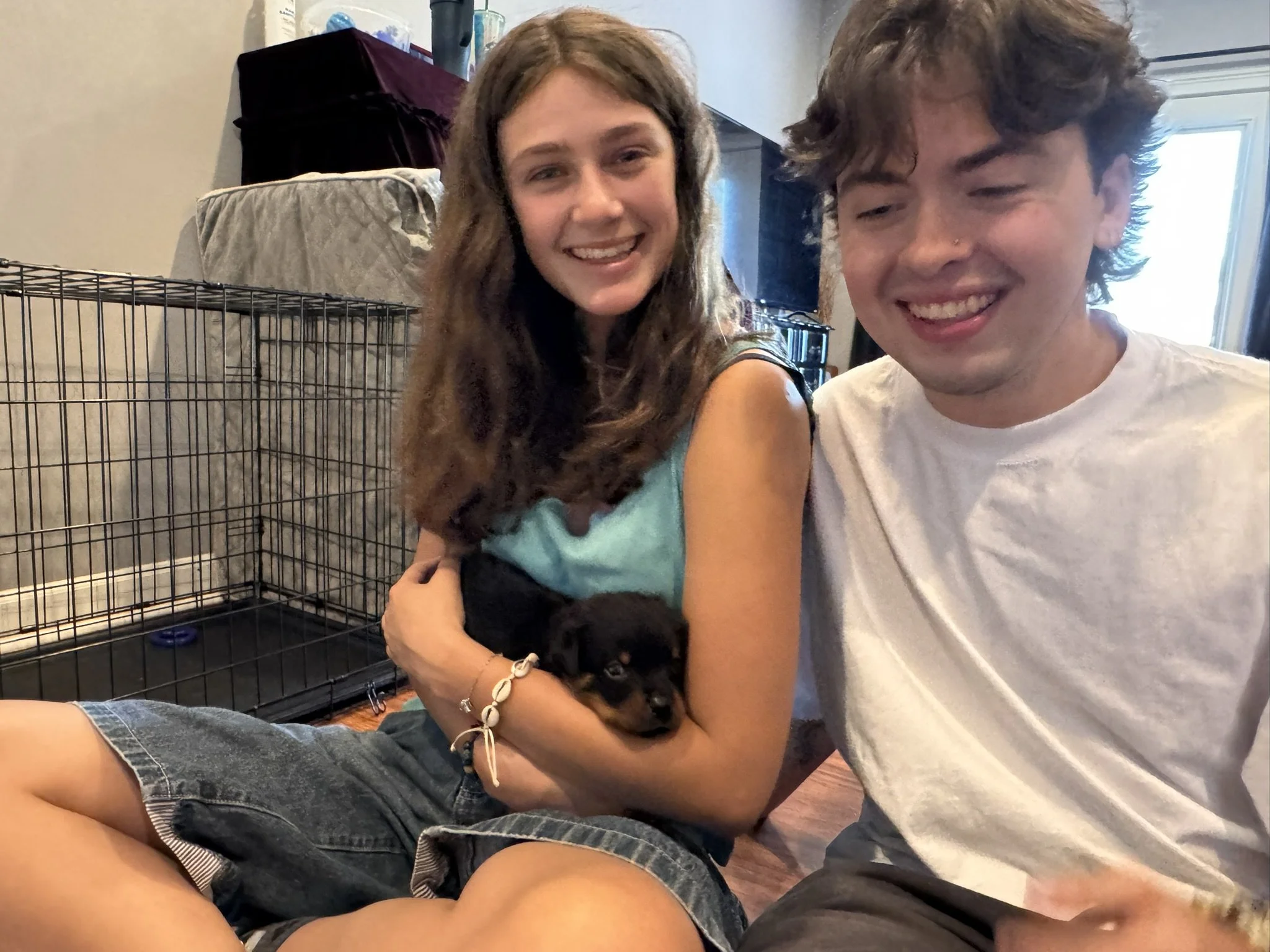 Two young people, a woman with long, wavy brown hair and a man with curly brown hair, smiling and sitting close together inside a house with a large pet cage and a window in the background.