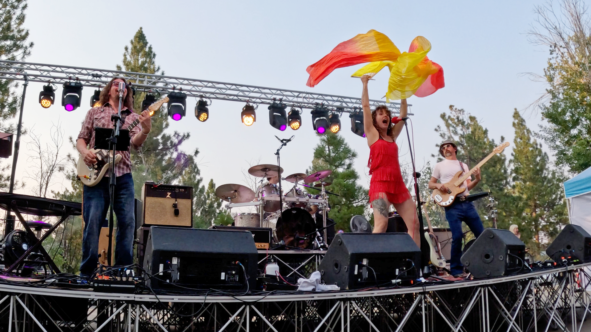 A band performs on an outdoor stage with purple lighting. The lead singer, wearing a red dress, is holding a yellow scarf and singing into a microphone. There are guitarists on either side, and a drummer in the background. Trees can be seen behind th