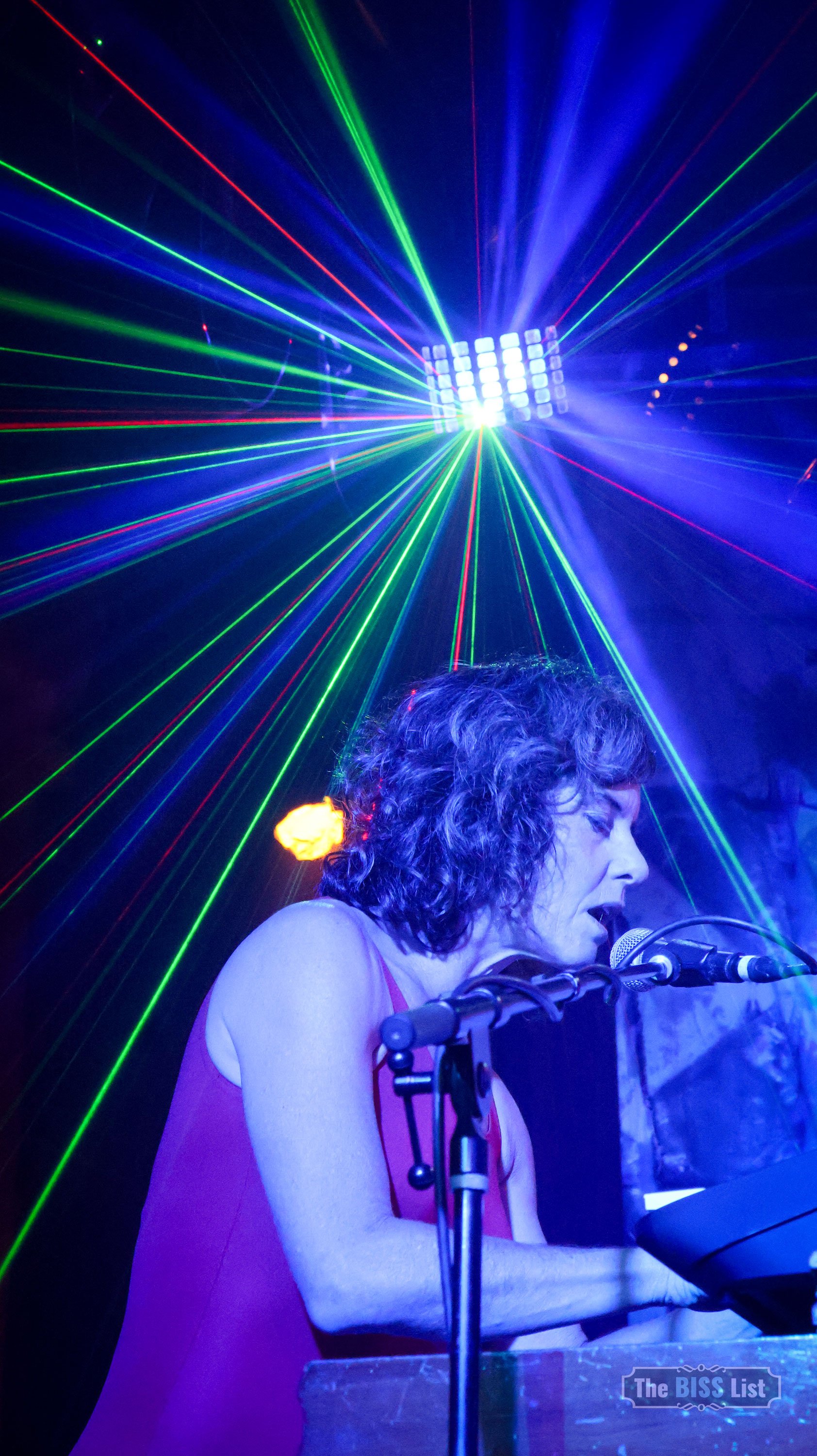 A woman with curly hair singing into a microphone while playing a keyboard, with colorful laser lights creating a pattern in the background.
