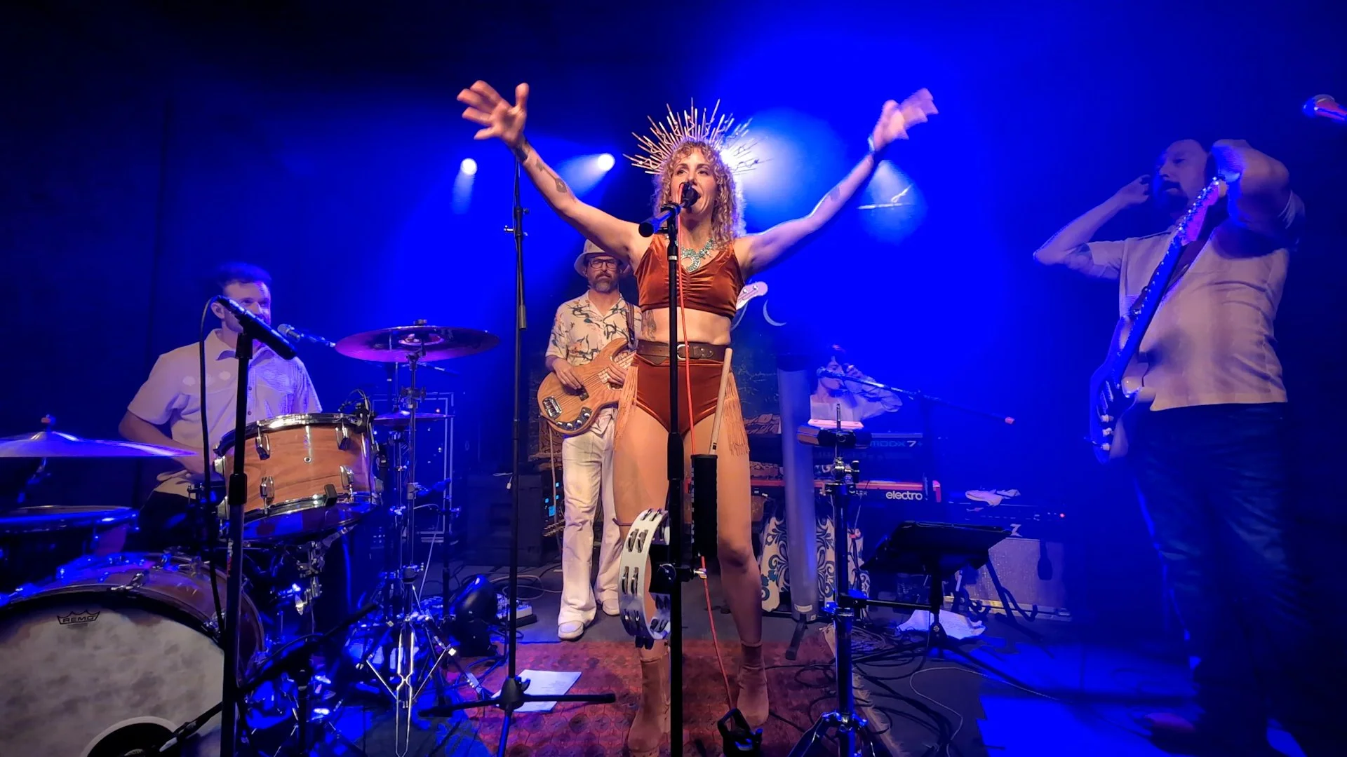 A band performs on stage with a female singer in the center wearing a metallic crown and matching outfit, surrounded by musicians playing drums, bass, guitar, and keyboard, with blue stage lighting.