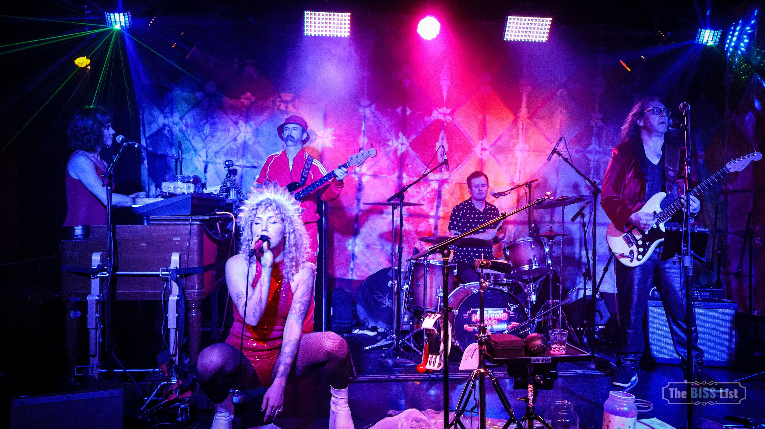 A musical band performing on stage with vibrant colorful lights, including a woman singing in the foreground, and four musicians playing instruments behind her.