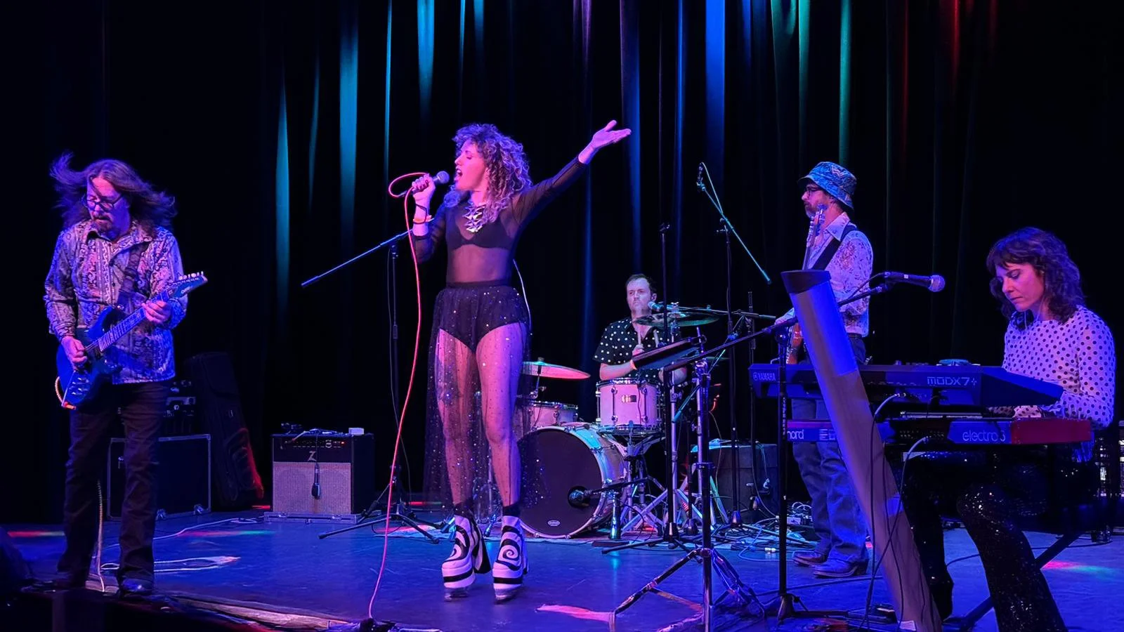 A band performing on stage with five members: guitarist, female lead singer, drummer, keyboardist, and saxophonist with microphones and musical equipment under colorful stage lighting.