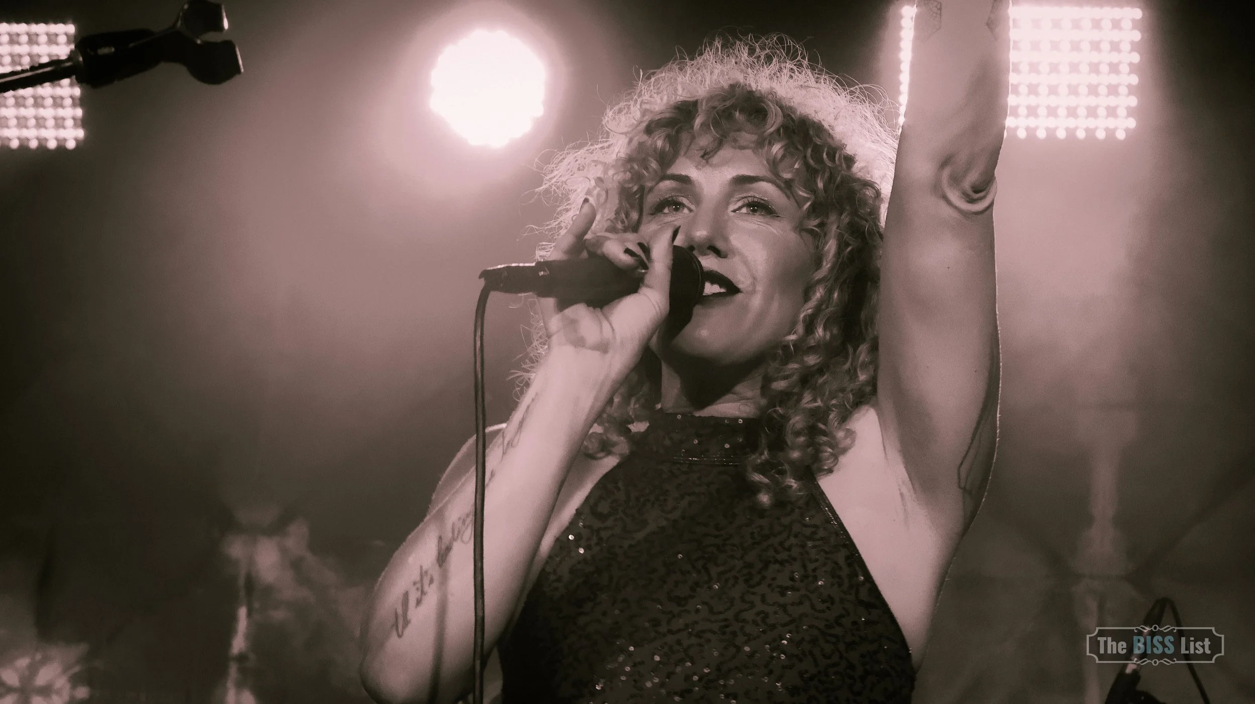 A woman with curly hair singing into a microphone on stage, with bright stage lights behind her.