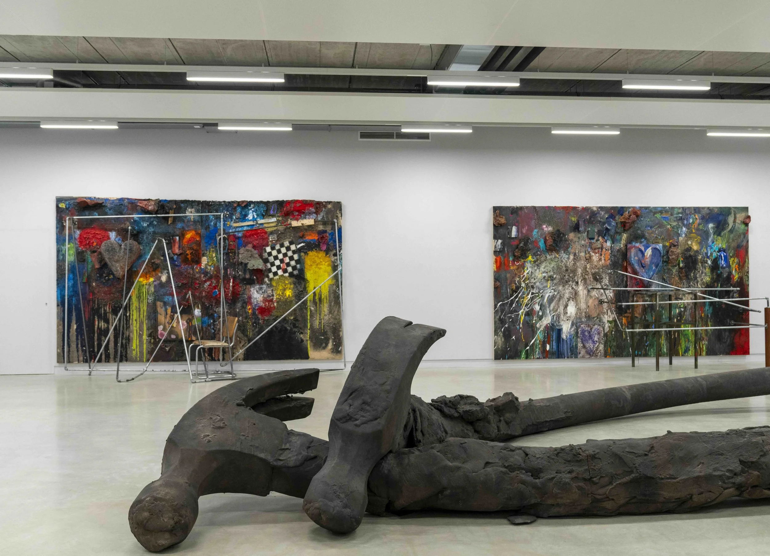 Oversized metal hammer sculpture lay on the floor of a gallery with colorful paintings in the background hanging on the wall