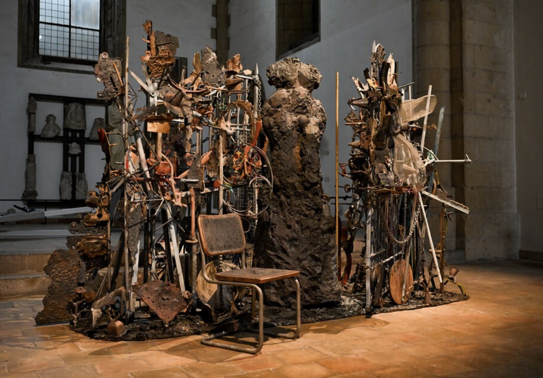 A sculpture installation by artist Jim Dine, comprised of clustered found objects like chairs, posts, and clay