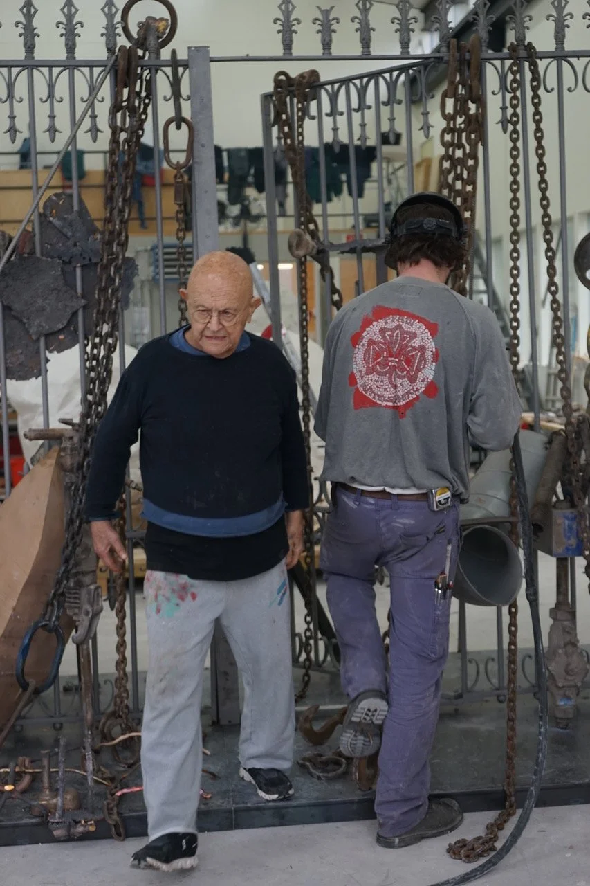 A candid photo of artist Jim Dine walking through a gate wearing paint covered clothes