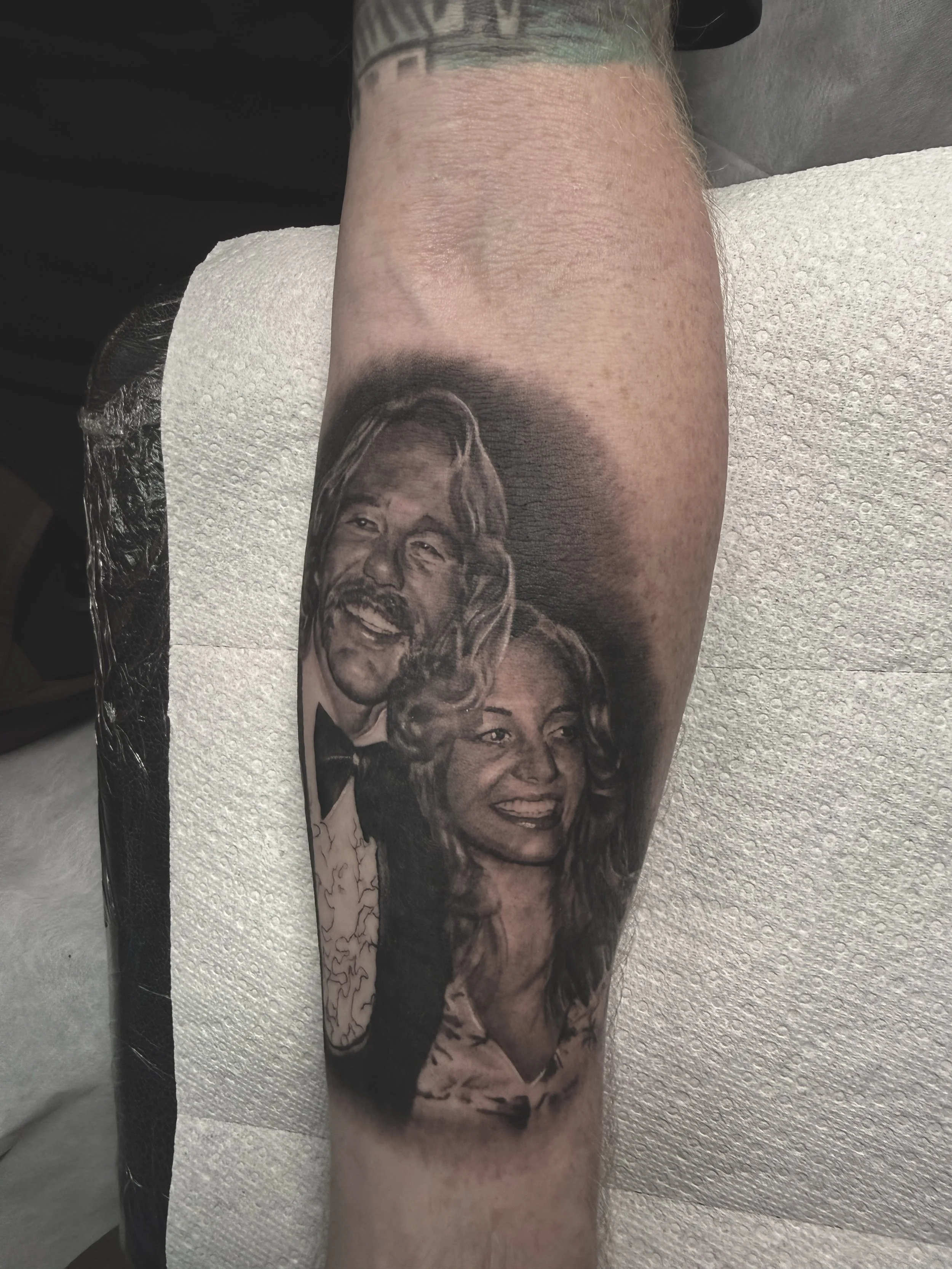Black and gray tattoo of a man with long hair and a beard, smiling in a tuxedo, and a woman with long hair and a smile, wearing a dress, on a person's arm done by Richy Reynolds at Harp and Rose Tattoo in Ventura California