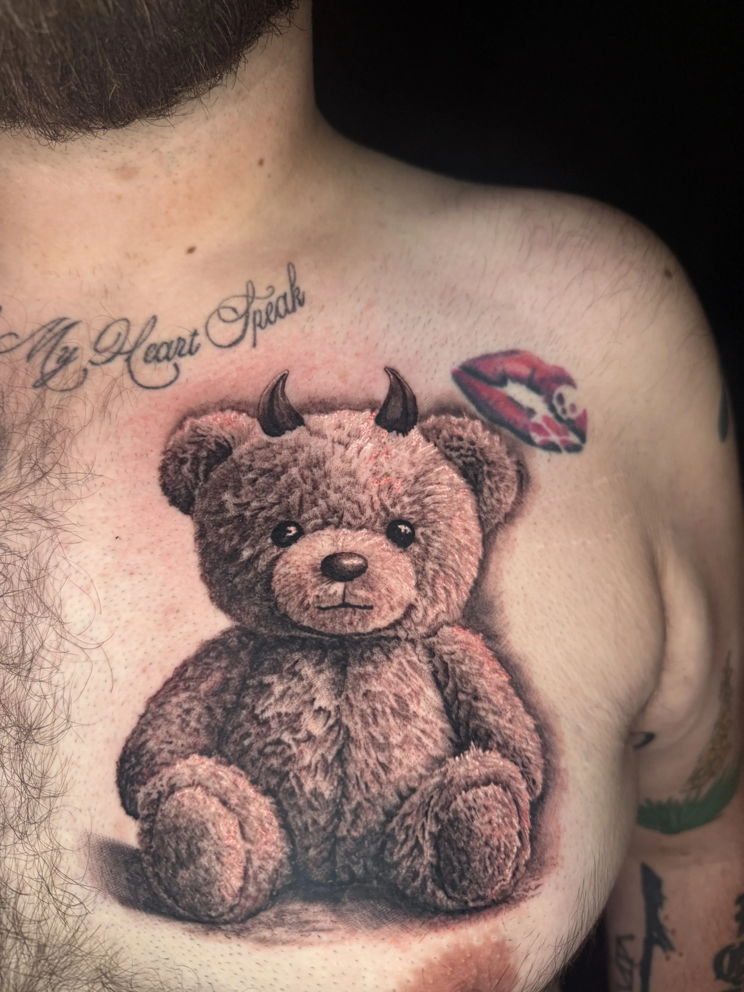 Teddy bear tattoo with black horns, next to text saying "My Heart Speaks" and a small, colorful lip tattoo above it done by Richy Reynolds at Harp and Rose Tattoo in Ventura California