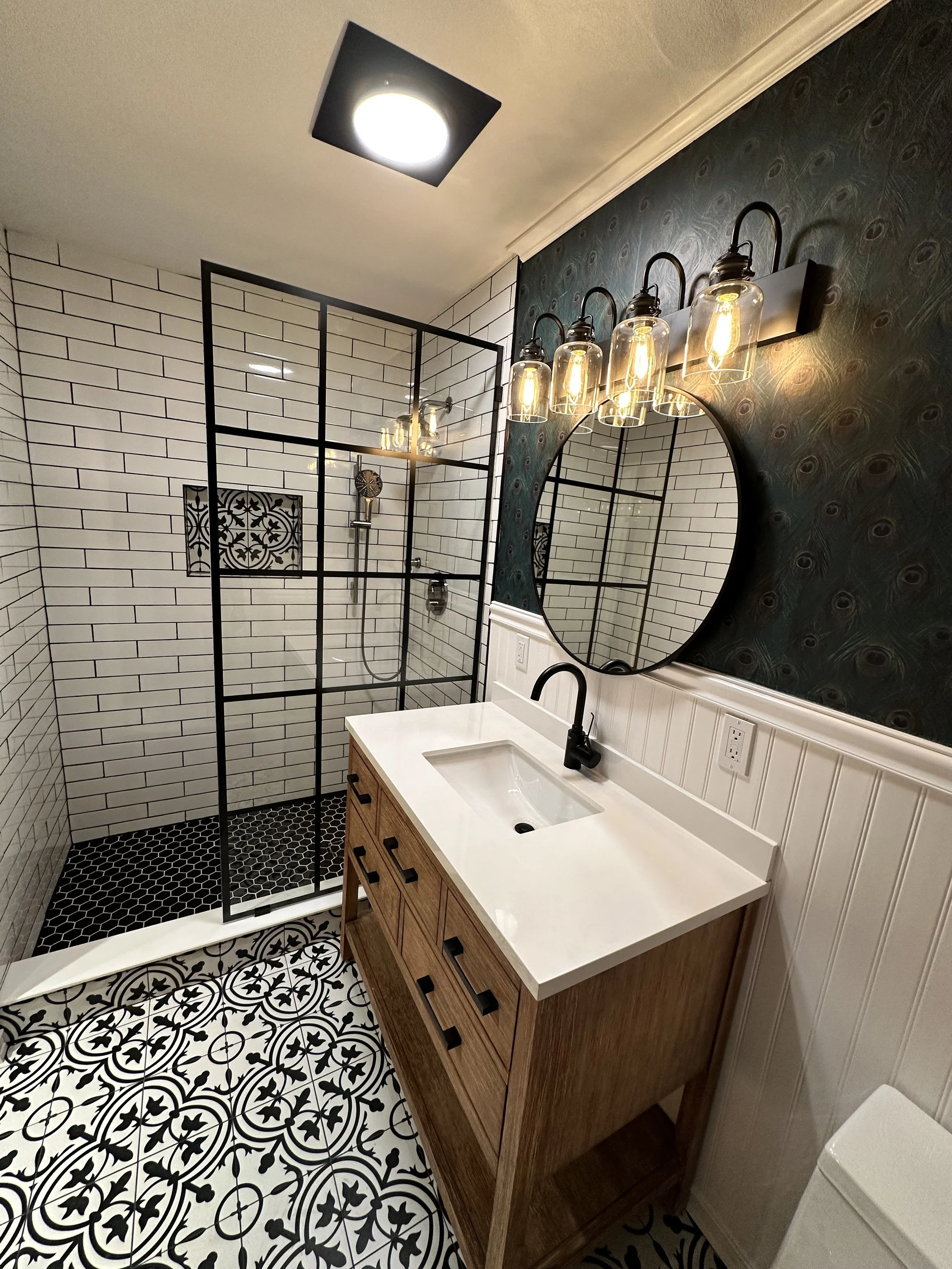 Modern bathroom with a walk-in shower with black metal framing, white subway tile walls, and black hexagonal floor tiles. There is a wooden vanity with a white countertop and a round mirror, illuminated by a multi-bulb light fixture. Partial view of 