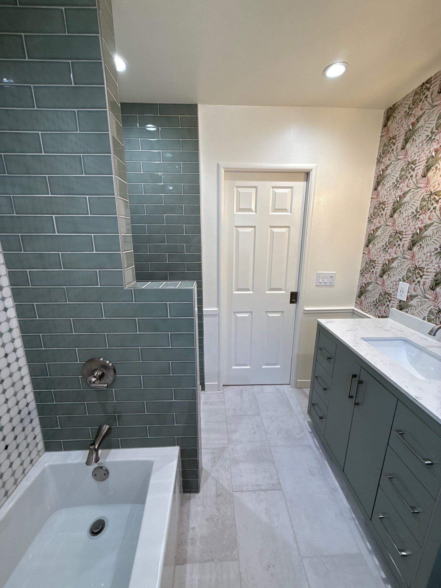 A bathroom with teal subway tile on the shower wall, a bathtub, a gray vanity with a white marble countertop, a floral wallpaper, and a white door.