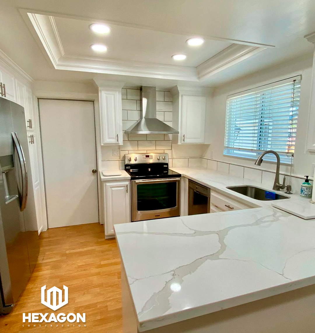 A modern kitchen with white cabinetry, a stainless steel stove and range hood, a side-by-side refrigerator, a window with blinds, and a white countertop with gray veining. The ceiling has recessed lighting and decorative crown molding, and there is a