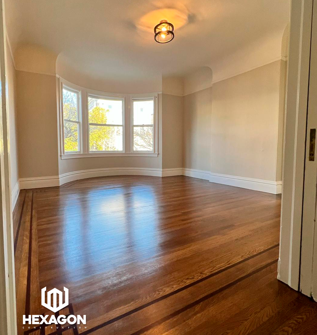 Empty room with hardwood floors, large bay window, beige walls, and a ceiling light fixture.