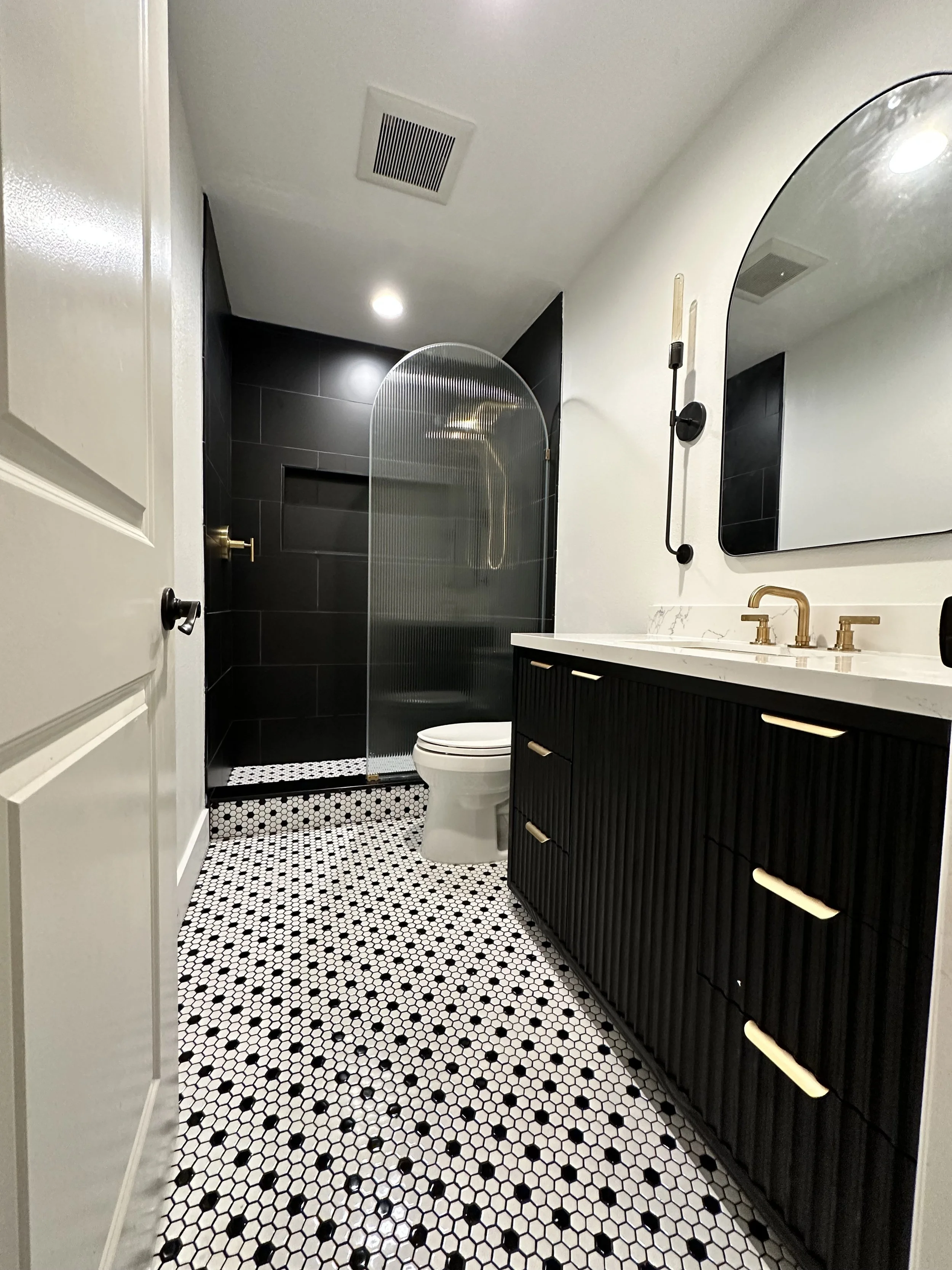 Modern bathroom with black and white mosaic tile floor, white walls, black vanity with brass handles and fixtures, large mirror, black walk-in shower with curved glass door, and a toilet.