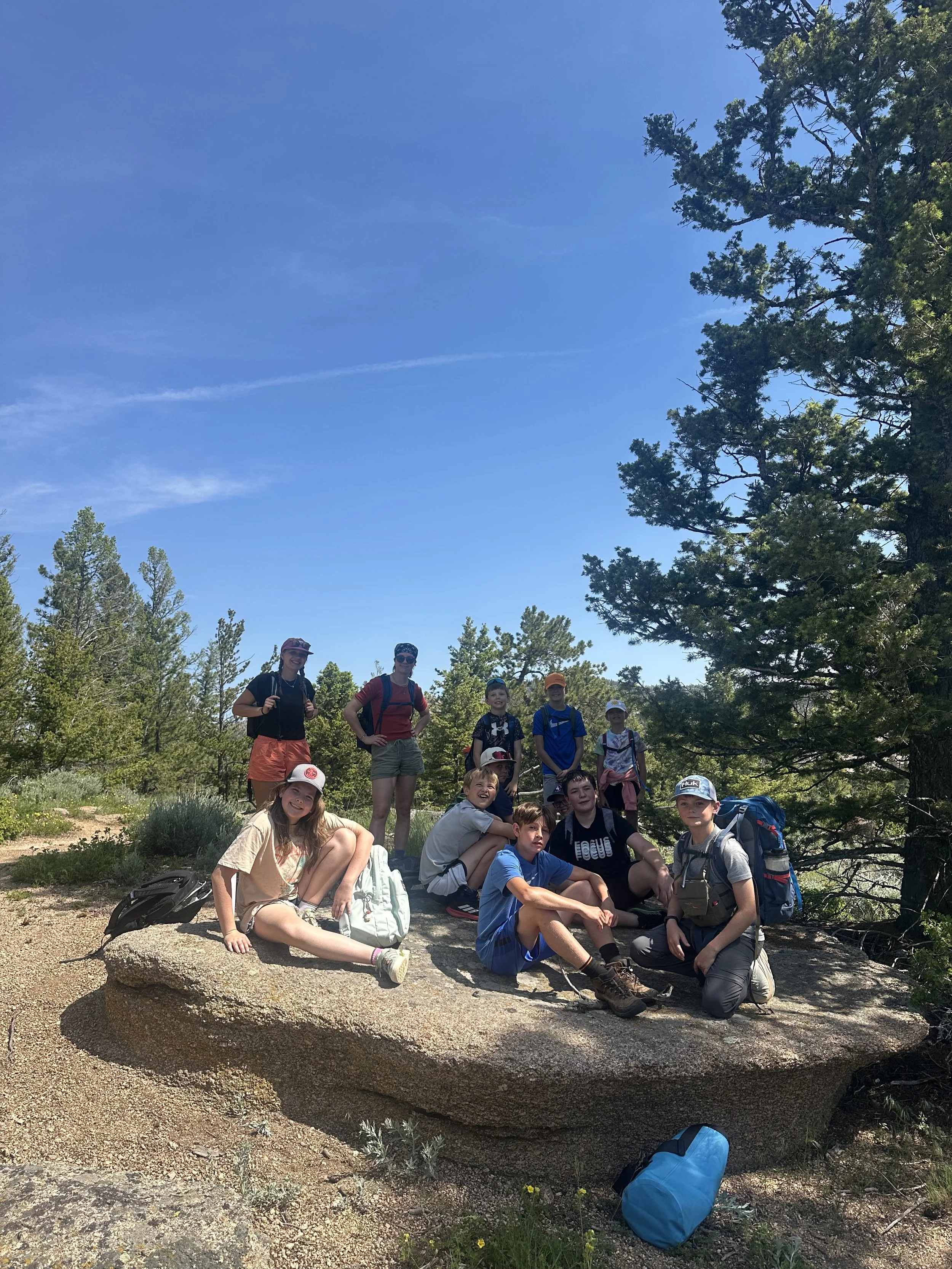 Group of children and teenagers on a large rock in a forested area, some with backpacks, enjoying a hike on a sunny day.