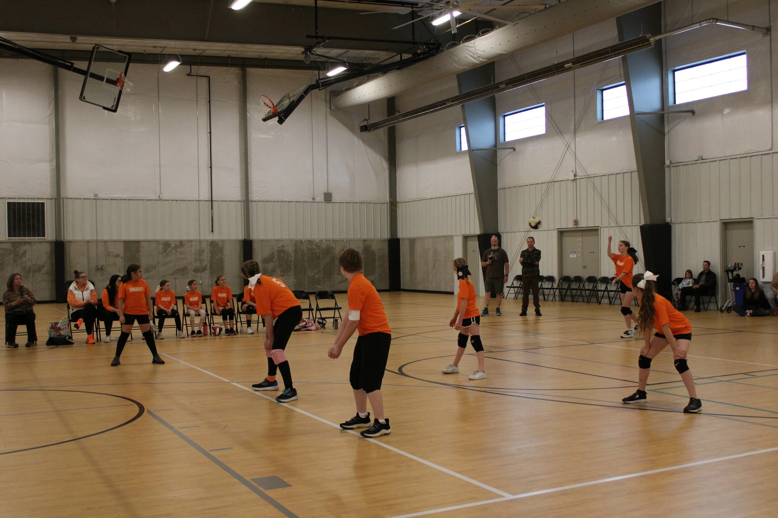Girls playing volleyball in an indoor gymnasium during a match, with a team seated on the bench and spectators watching in the background.