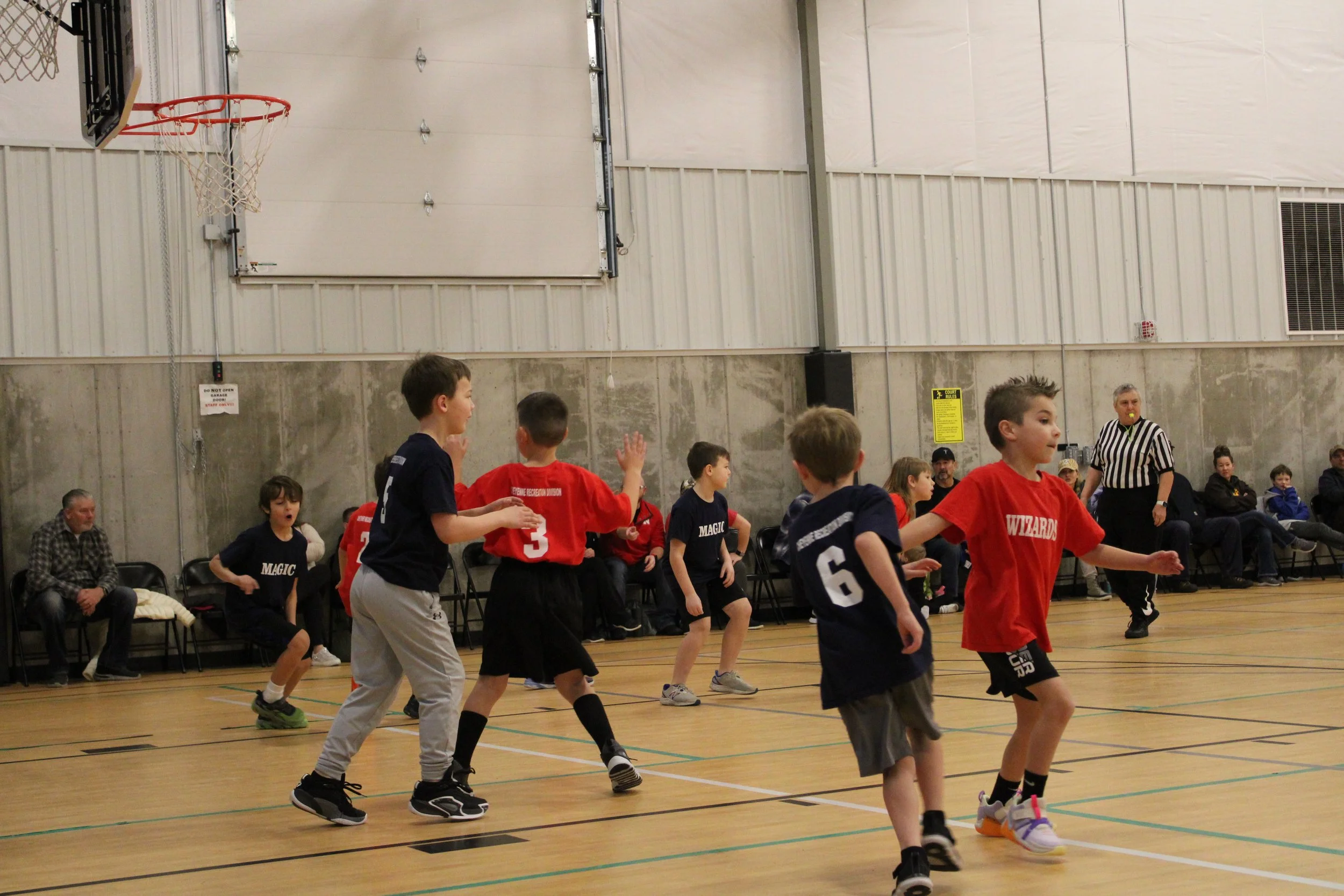Young children playing basketball in an indoor gym, with spectators seated along the wall.
